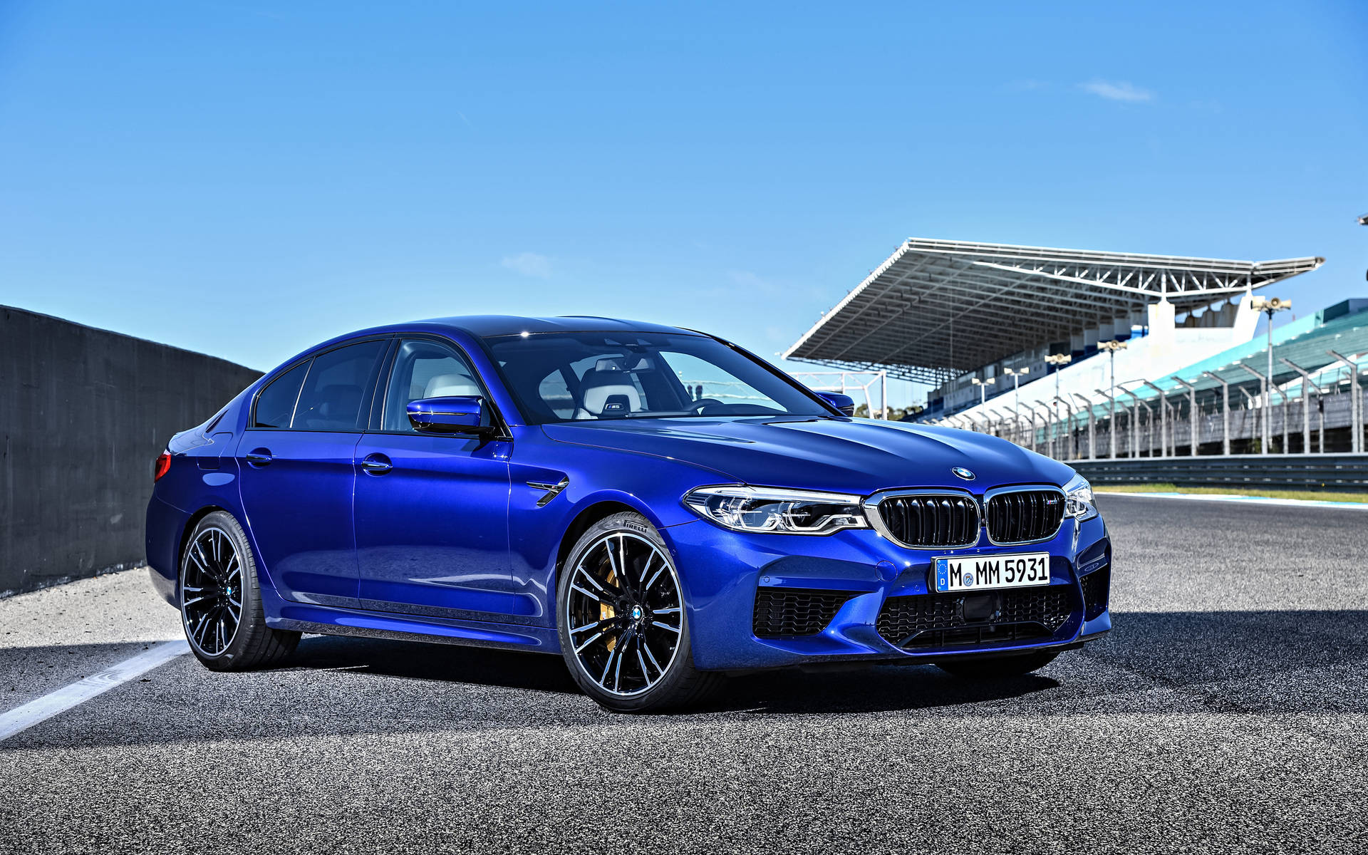 Bmw 桌布 壁紙 — 一輛鮮藍色BMW M5房車在明亮的陽光下，在瀝青賽道上閃閃發光，背景有看台。