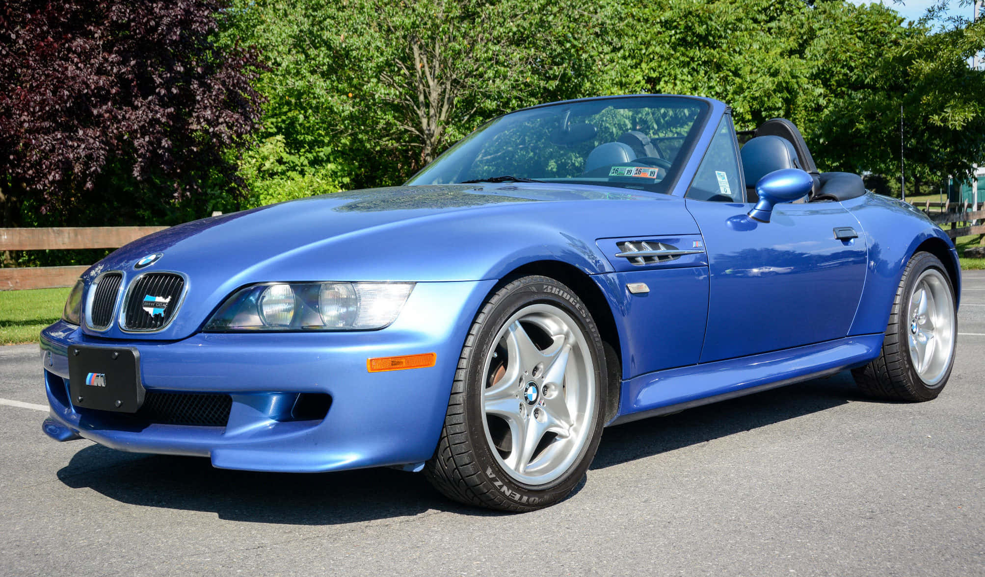 Bmw Z3 桌布 壁紙 — 一輛鮮藍色BMW Z8開篷車停在瀝青路上，背景有茂密的綠樹，在明亮的日光下。