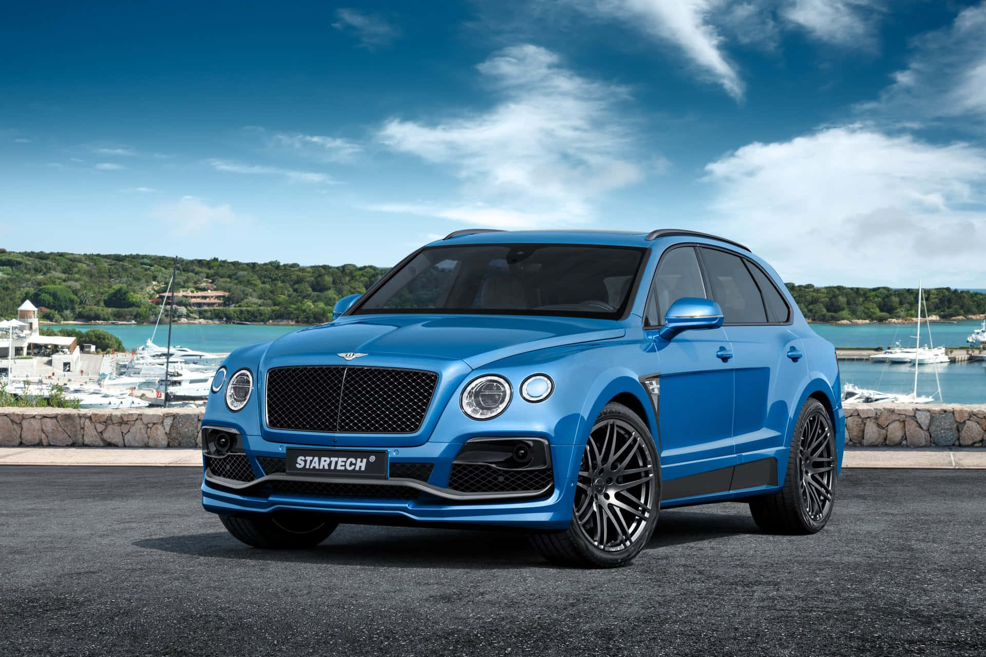 Bentley Bentayga 桌布 壁紙 — 一輛鮮藍色Startech改裝的賓利Bentayga SUV停泊在沿海道路上，背景是碼頭和綠色山丘，天空部分多雲。