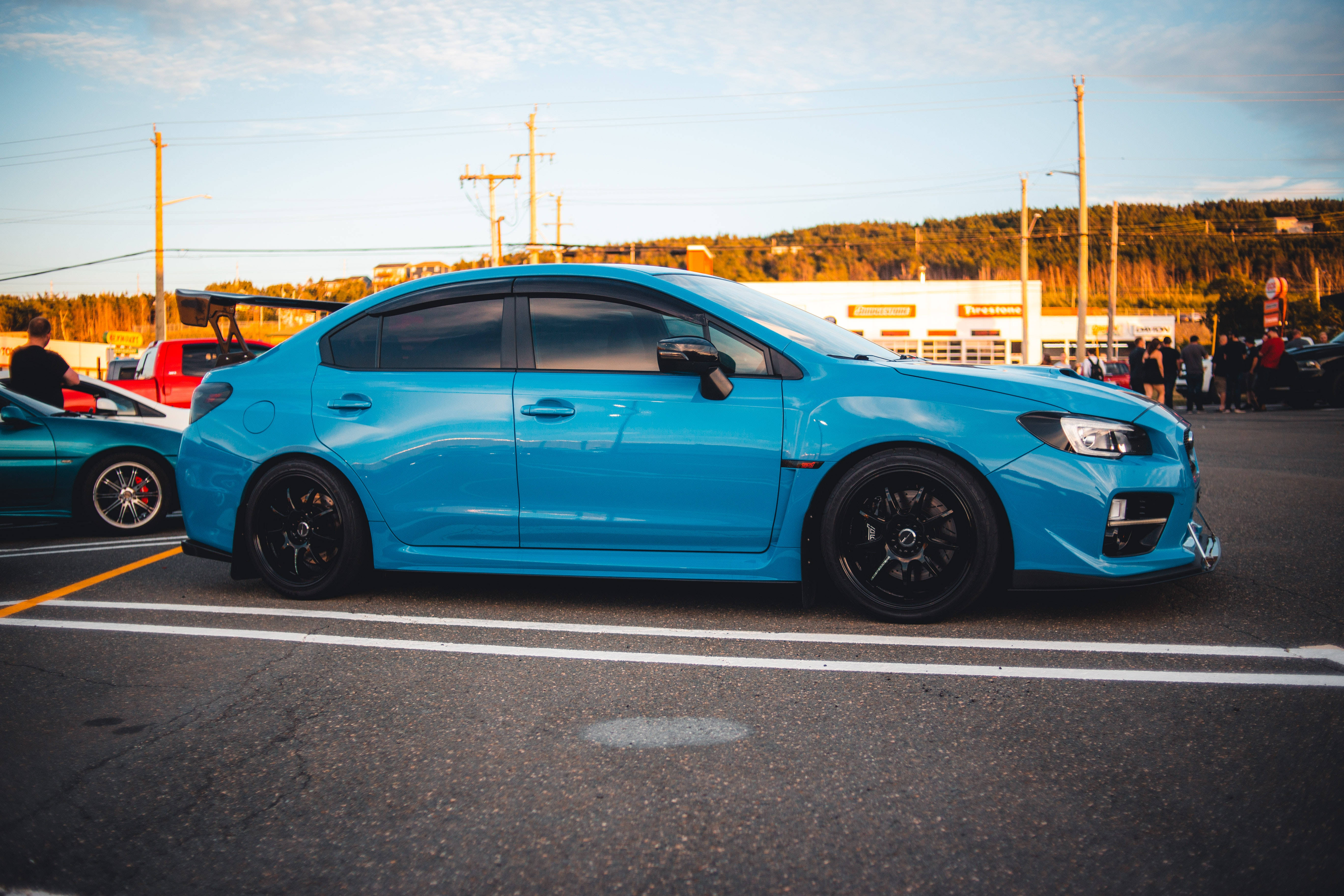 subaru 桌布 壁紙 — 一輛鮮藍色改裝房車配黑色輪圈，在黃金時段停泊在停車場，背景有人和樹木。