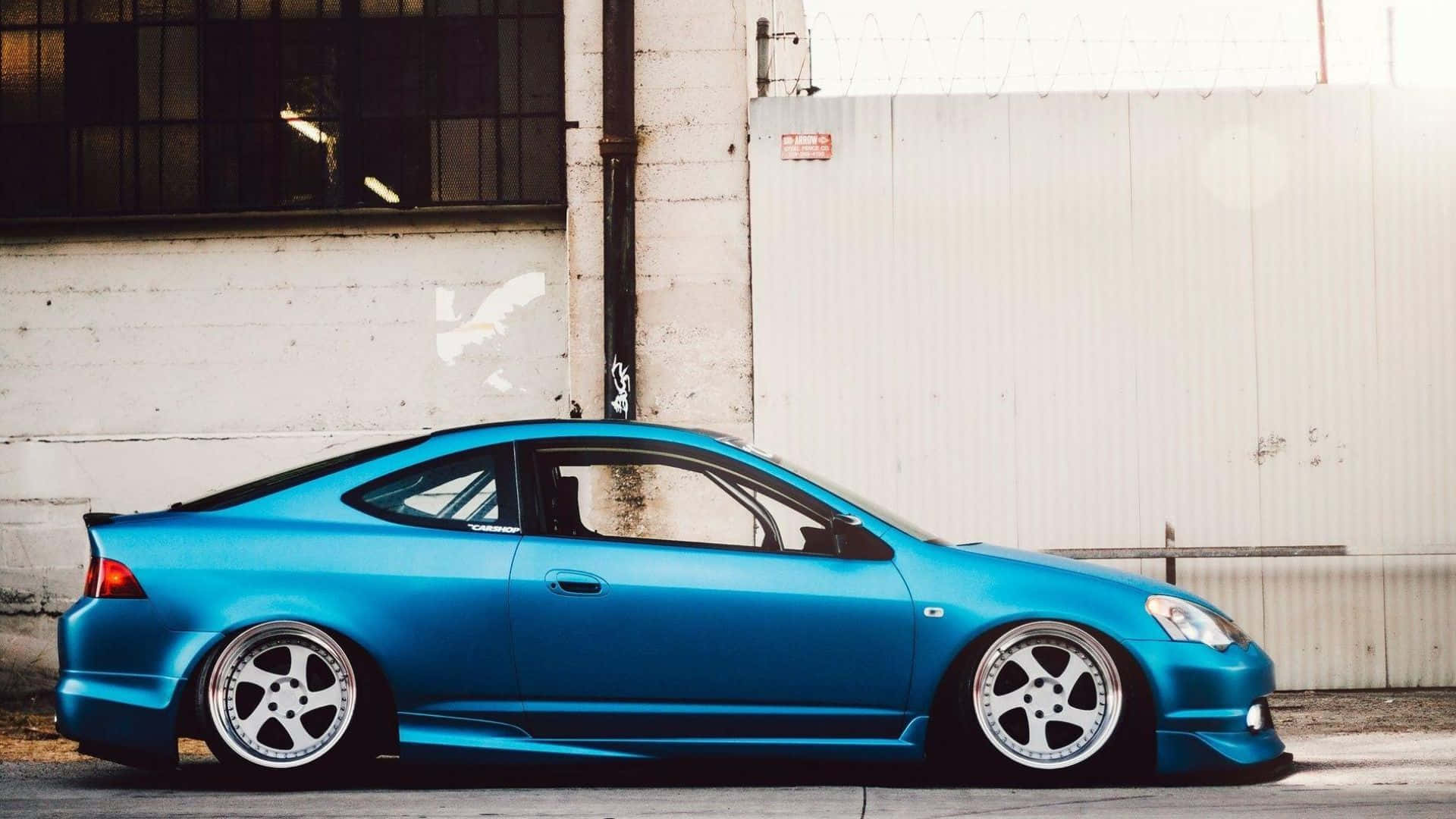 Rsx Type-s 桌布 壁紙 — 一輛鮮藍色改裝轎跑車，配備定制輪圈，停泊在風化的工業牆旁。
