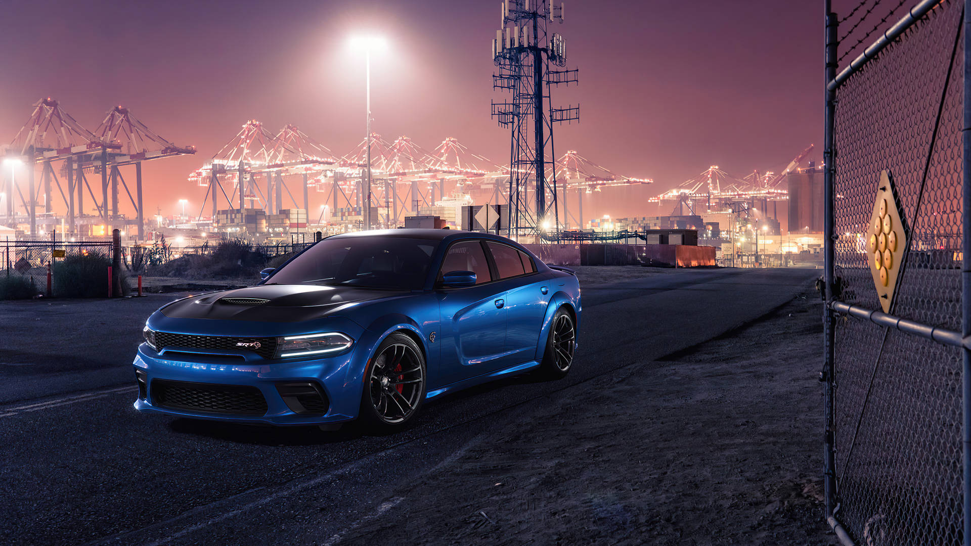 dodge challenger 桌布 壁紙 — 一輛鮮藍色的 Dodge Charger SRT Hellcat 停泊在黃昏時分的圍欄工業港口，背景有發光的起重機。