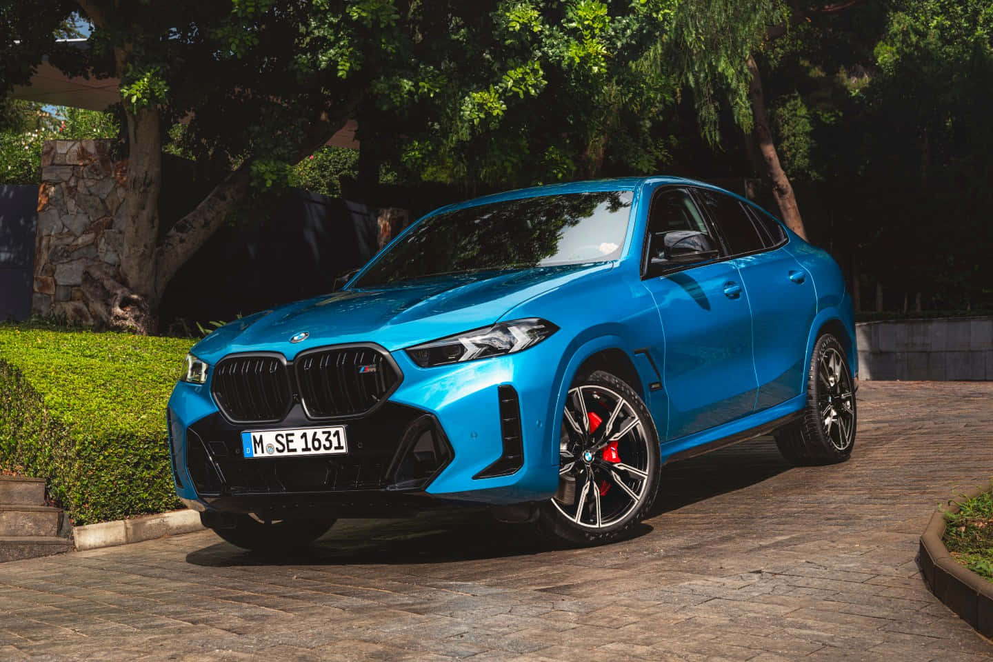 Bmw X5 桌布 壁紙 — 一輛鮮藍色的BMW X6 M停泊在石頭車道上，周圍環繞著茂密的綠樹和修剪整齊的樹籬。