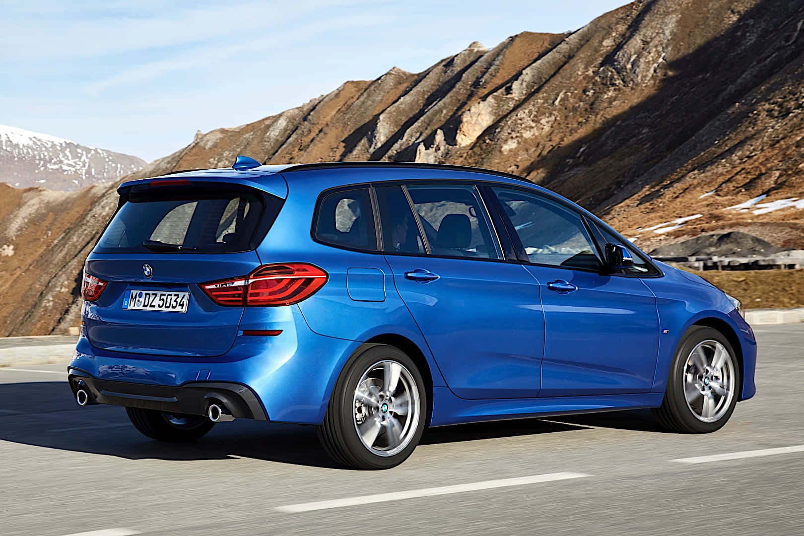 Bmw 2 Series 桌布 壁紙 — 一輛鮮藍色的BMW旅行車在晴朗的藍天下，沿著一條有崎嶇棕色山峰和積雪山頂的山路行駛。
