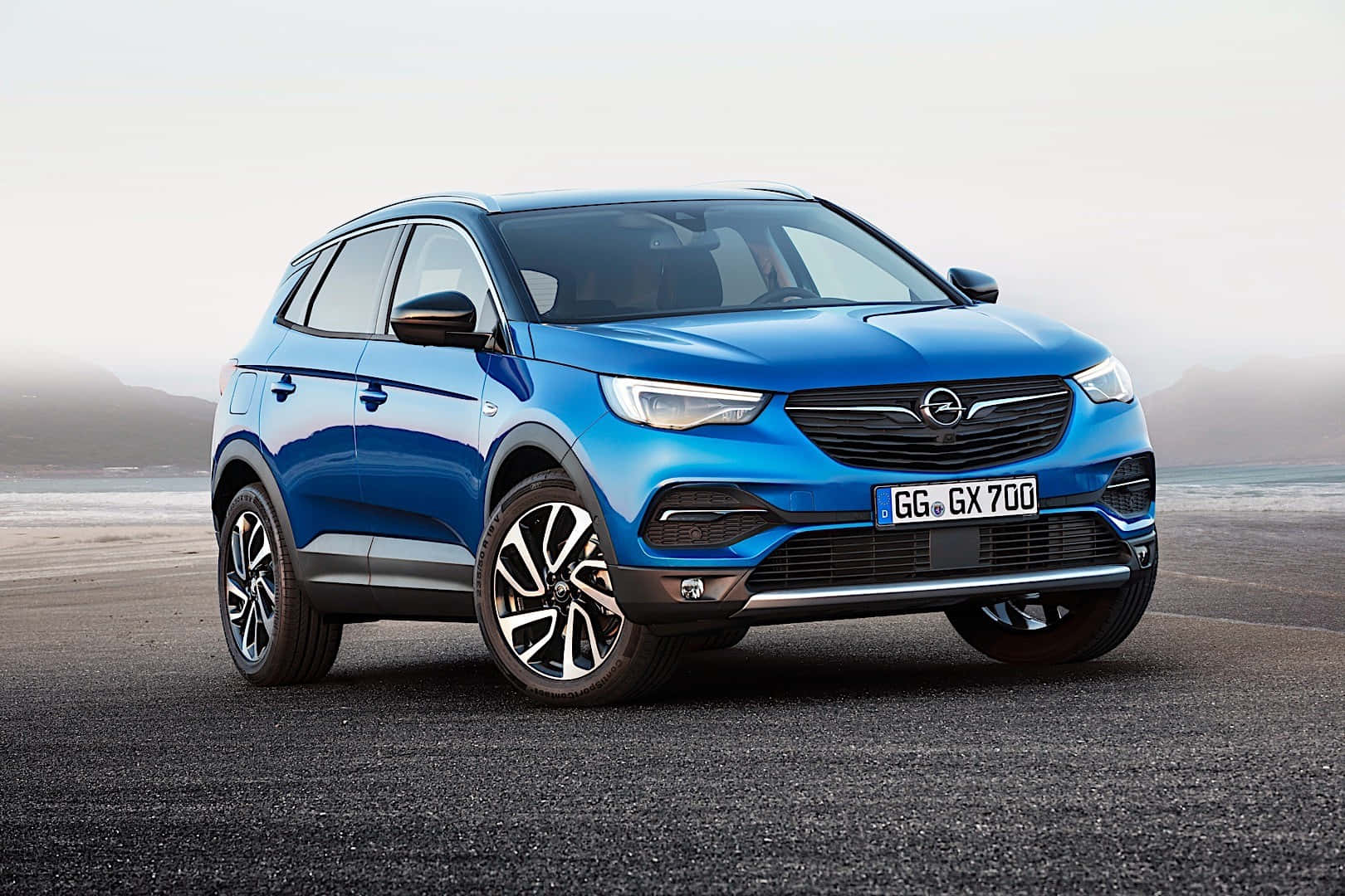 Opel Grandland X 桌布 壁紙 — 一輛鮮藍色的Opel SUV停泊在霧氣瀰漫的海岸公路上，遠山在柔和陰天之下。