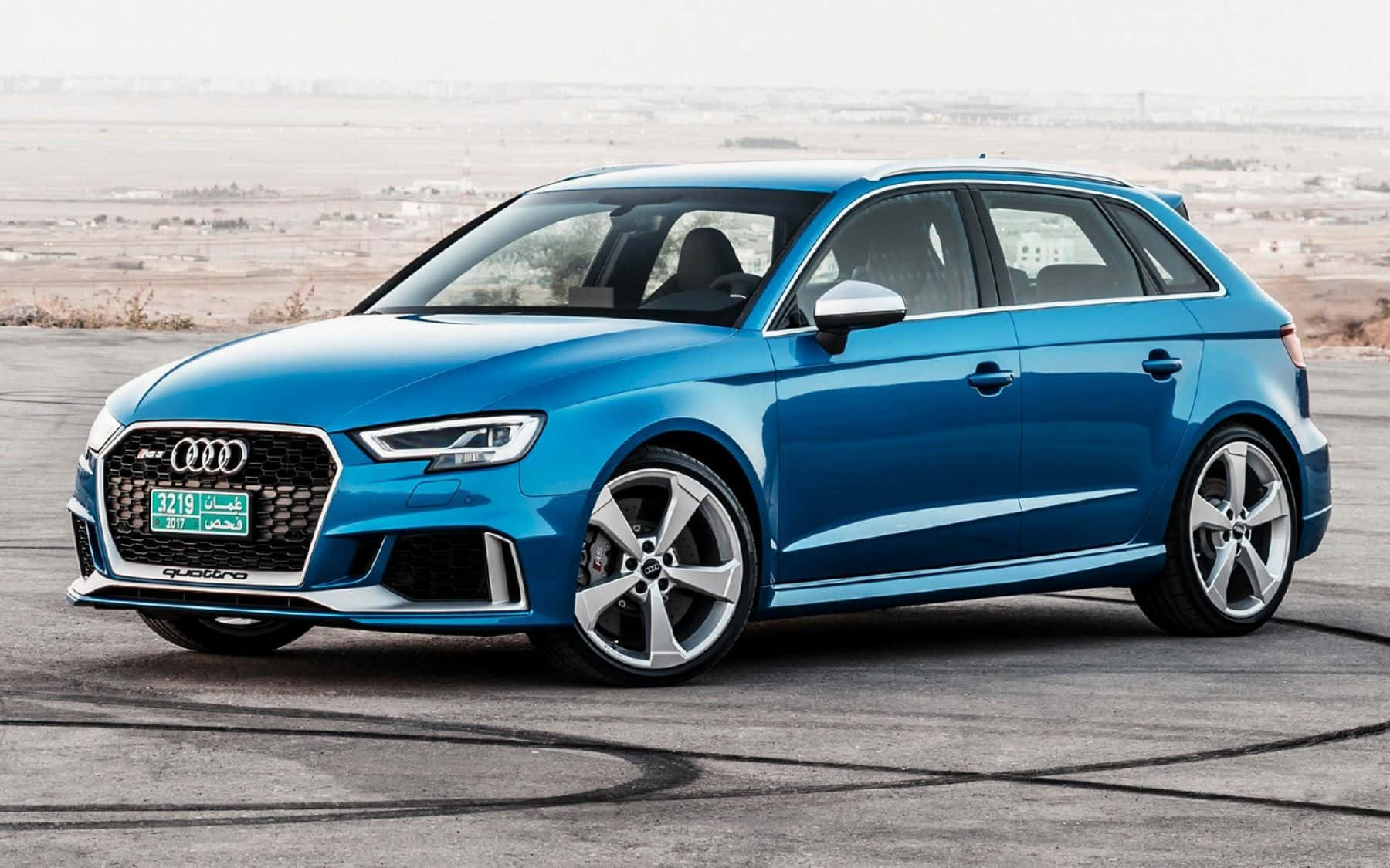 Audi Rs3 桌布 壁紙 — 一輛鮮藍色的奧迪RS3 Sportback停泊在鋪砌的停車場上，背景是朦朧的沙漠景觀。