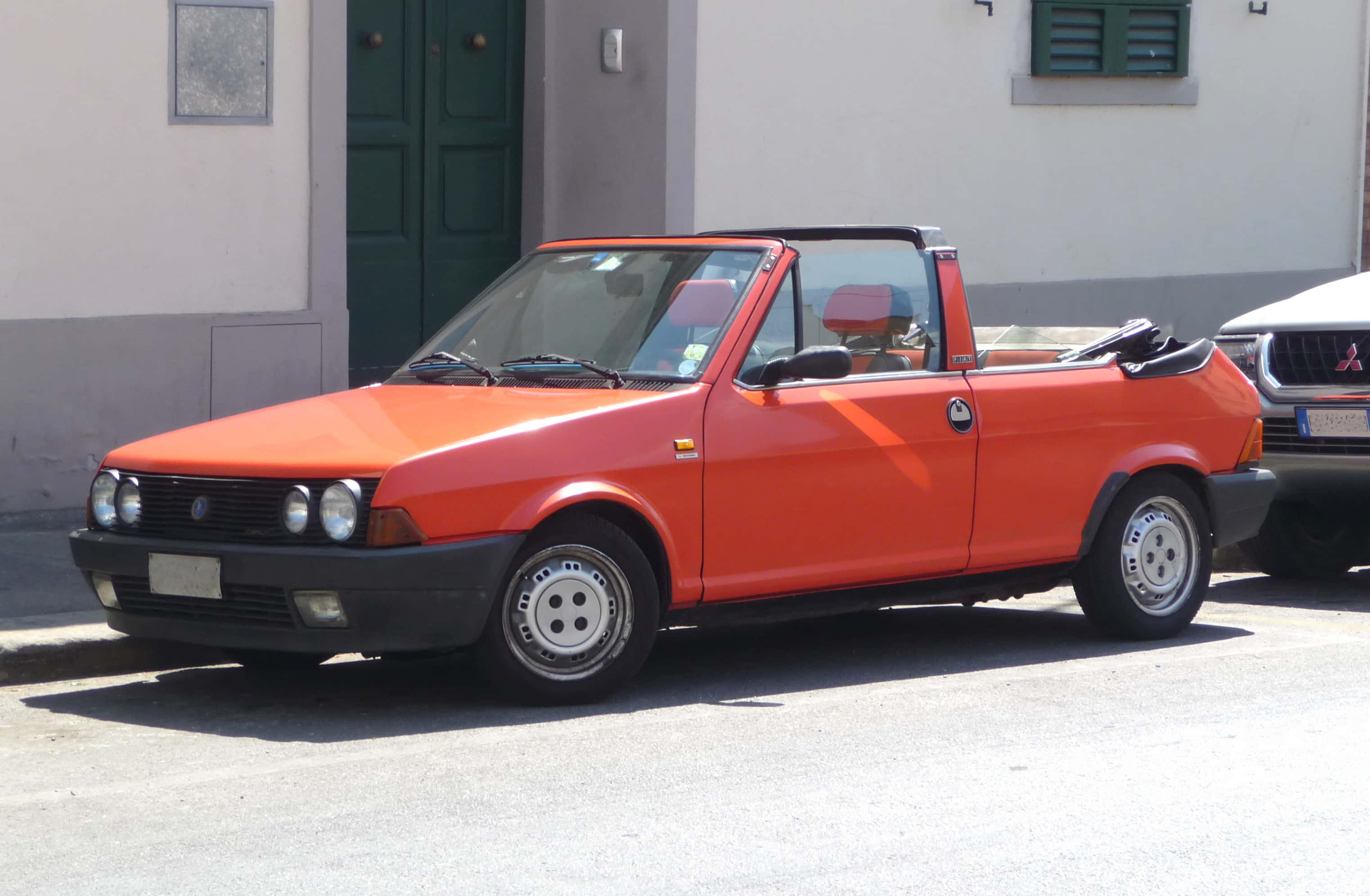 Fiat Ritmo 桌布 壁紙 — 一輛鮮豔的橙色Fiat 126 Spider開篷車停泊在陽光明媚的街道上，旁邊是一棟有綠色百葉窗的建築物。