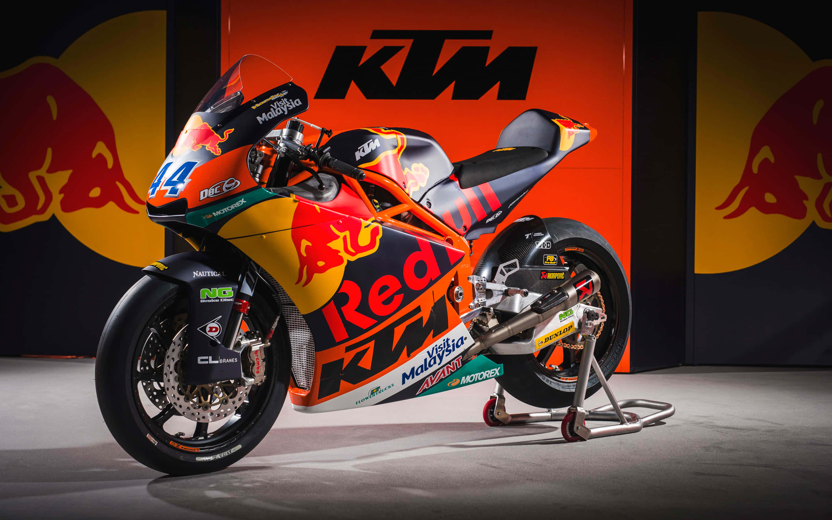 Ktm Bike 桌布 壁紙 — 一輛鮮豔的橙色和黑色KTM MotoGP賽車，帶有Red Bull品牌標誌，立在支架上，背景為品牌背景。