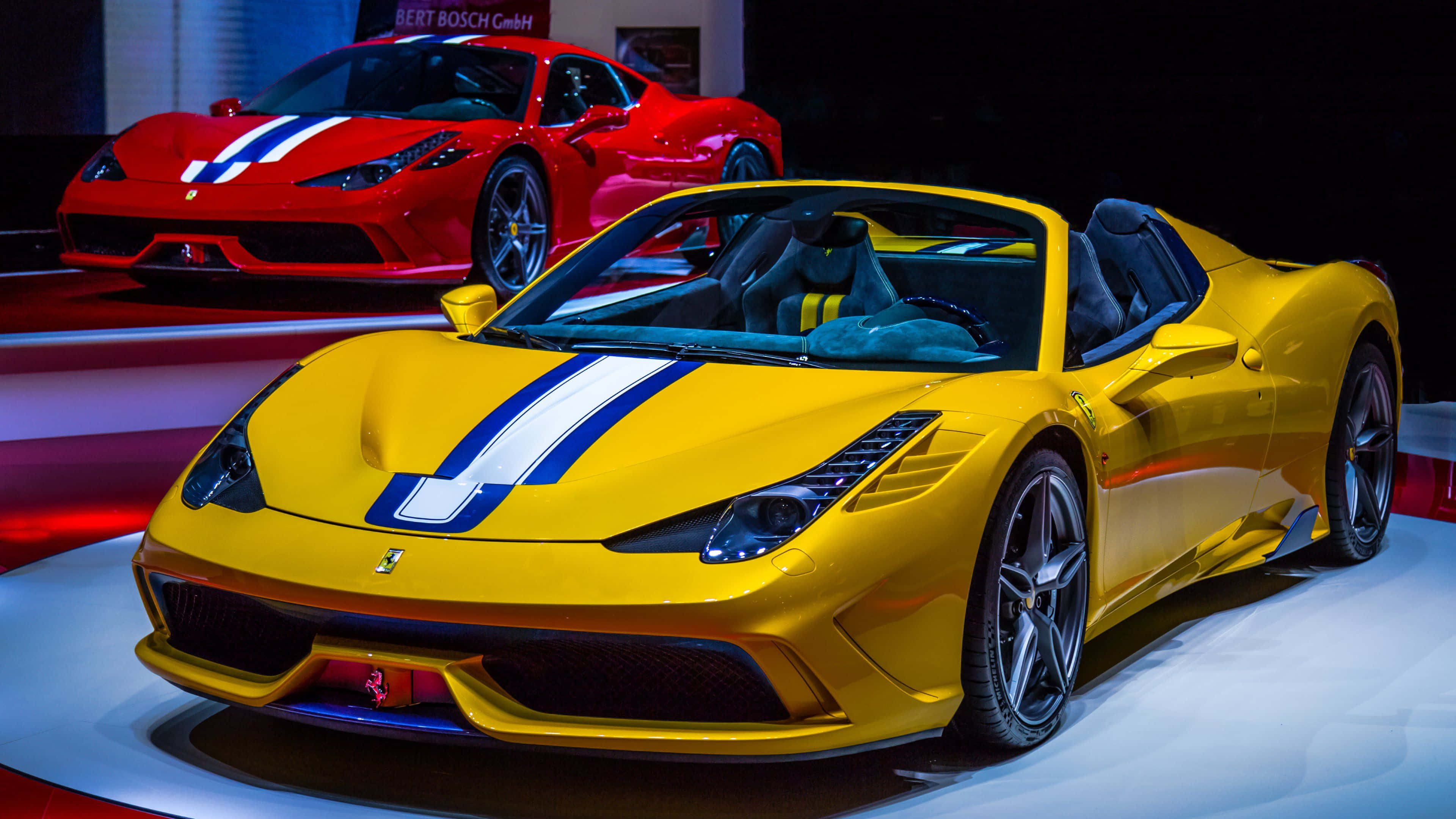Ferrari 458 Speciale 桌布 壁紙 — 一輛鮮黃色法拉利開篷跑車，帶有藍白賽車條紋，在明亮燈光下閃閃發光，停在一輛紅色法拉利前面，於車展中展出。