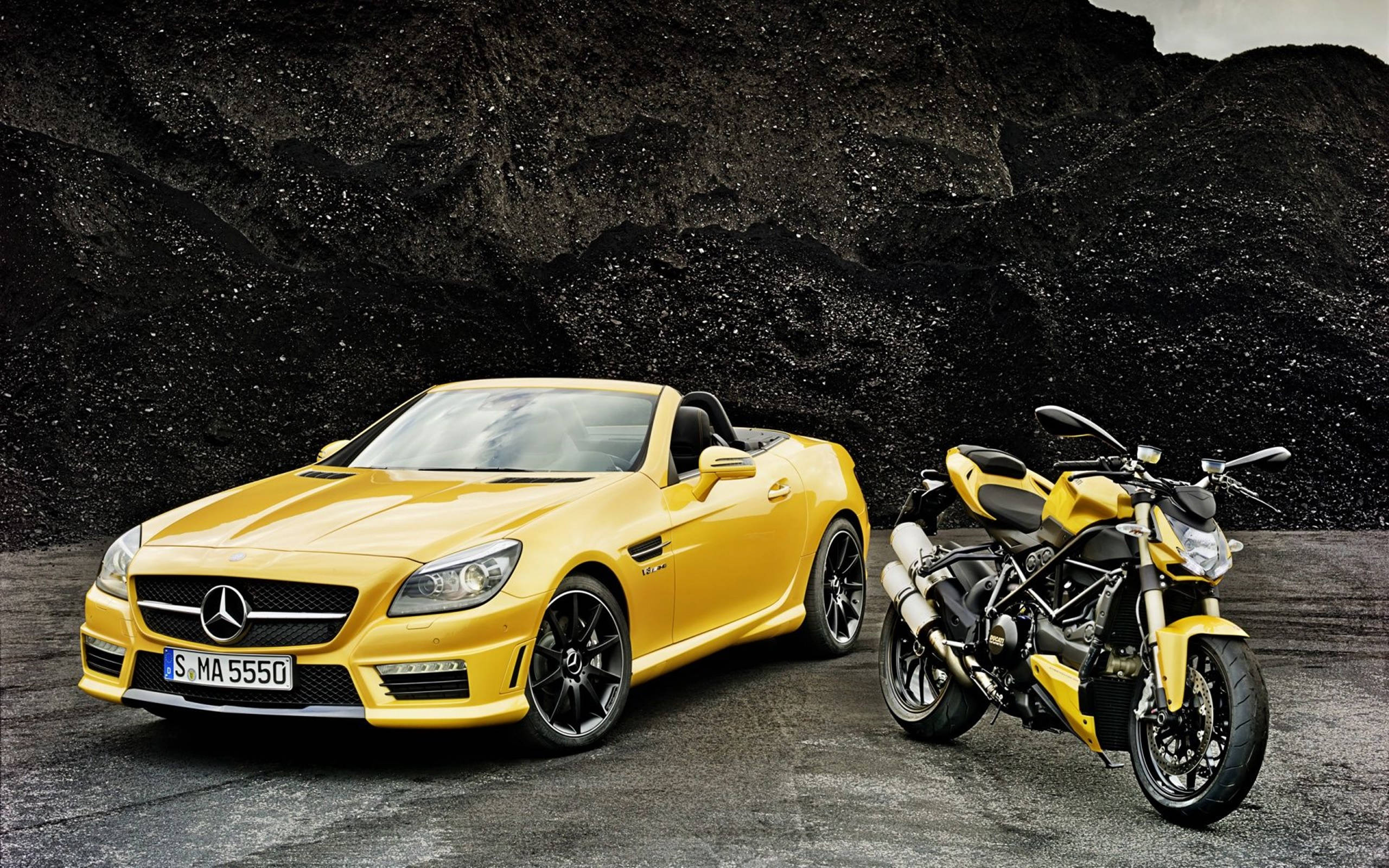 HD 桌布 壁紙 — 一輛鮮黃色的 Mercedes SLK 開篷車和一輛相配的黃色 Ducati 電單車並排停放在深色紋理岩石牆前。