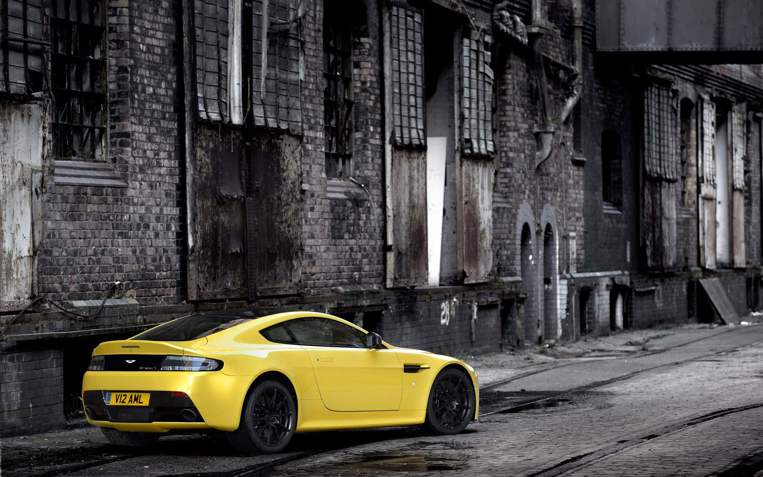 Aston Martin V12 Vantage 桌布 壁紙 — 一輛鮮黃色的Aston Martin跑車停泊在破舊磚倉庫旁一條佈滿灰塵的都市街道上。