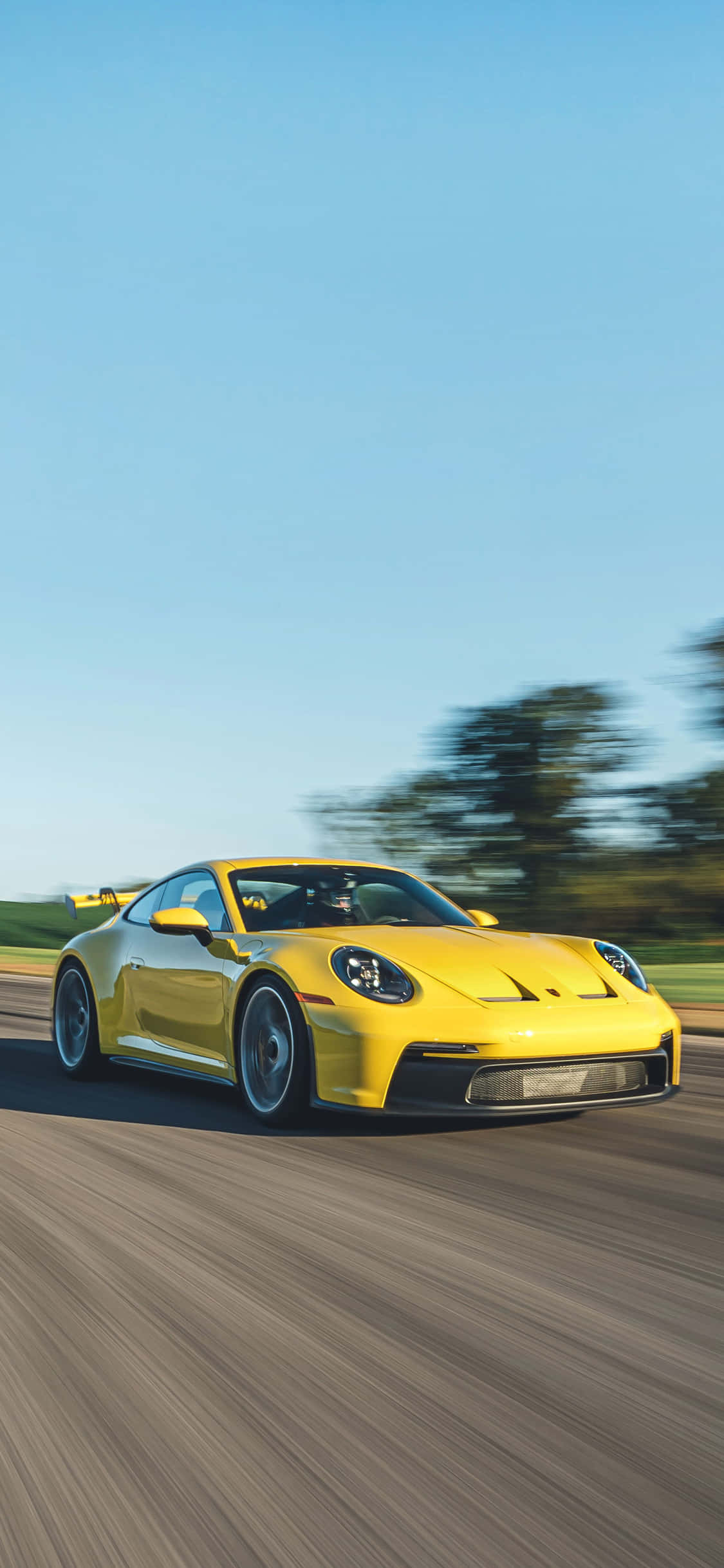 Porsche iPhone 桌布 壁紙 — 一輛鮮黃色的保時捷911 GT3 RS在陽光明媚的道路上疾馳，背景是模糊的綠樹和藍天。