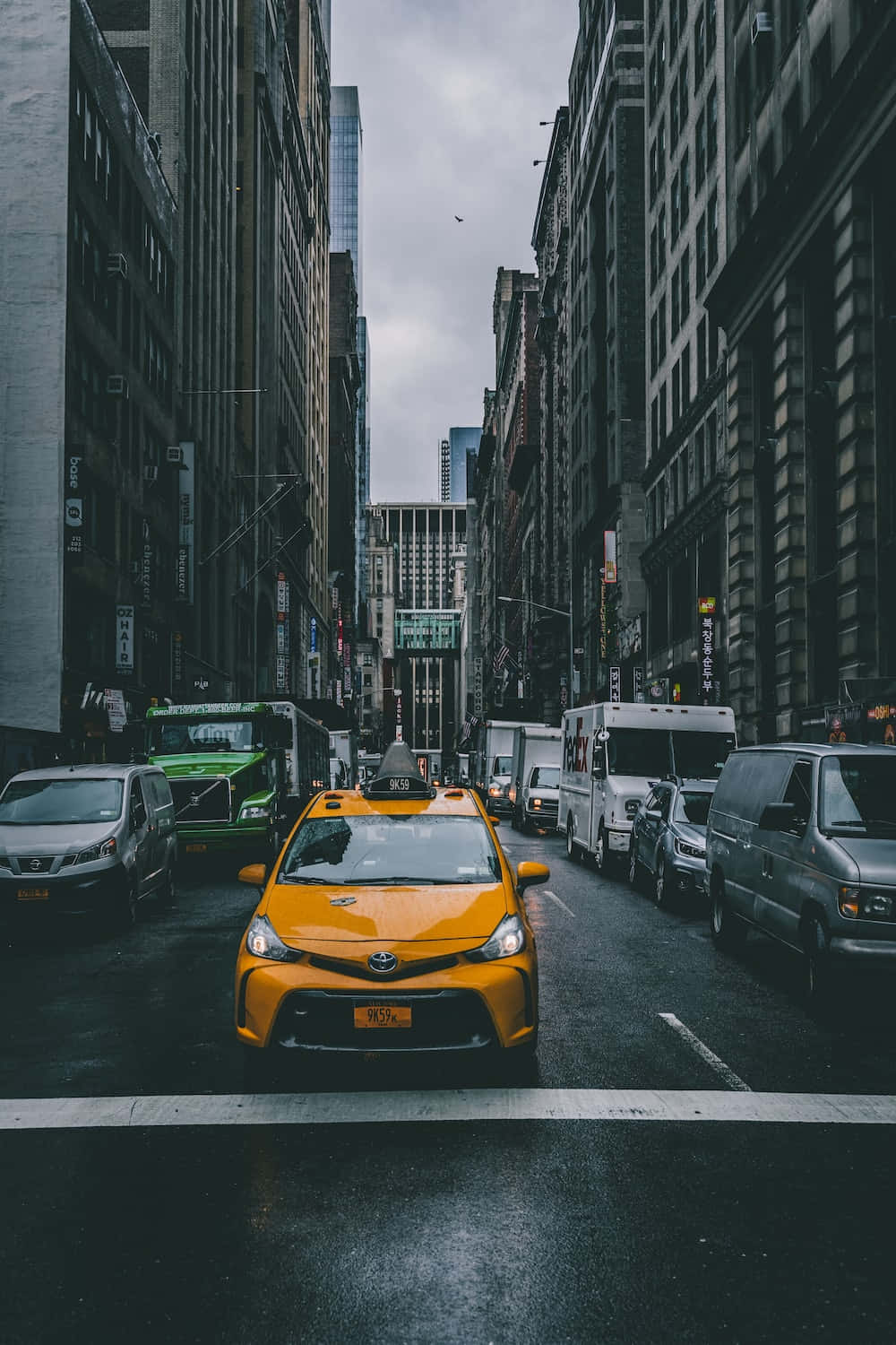 Yellow Cab 桌布 壁紙 — 一輛鮮黃色的士在陰天之下，沿著兩旁高聳摩天大樓的紐約市濕滑街道行駛。