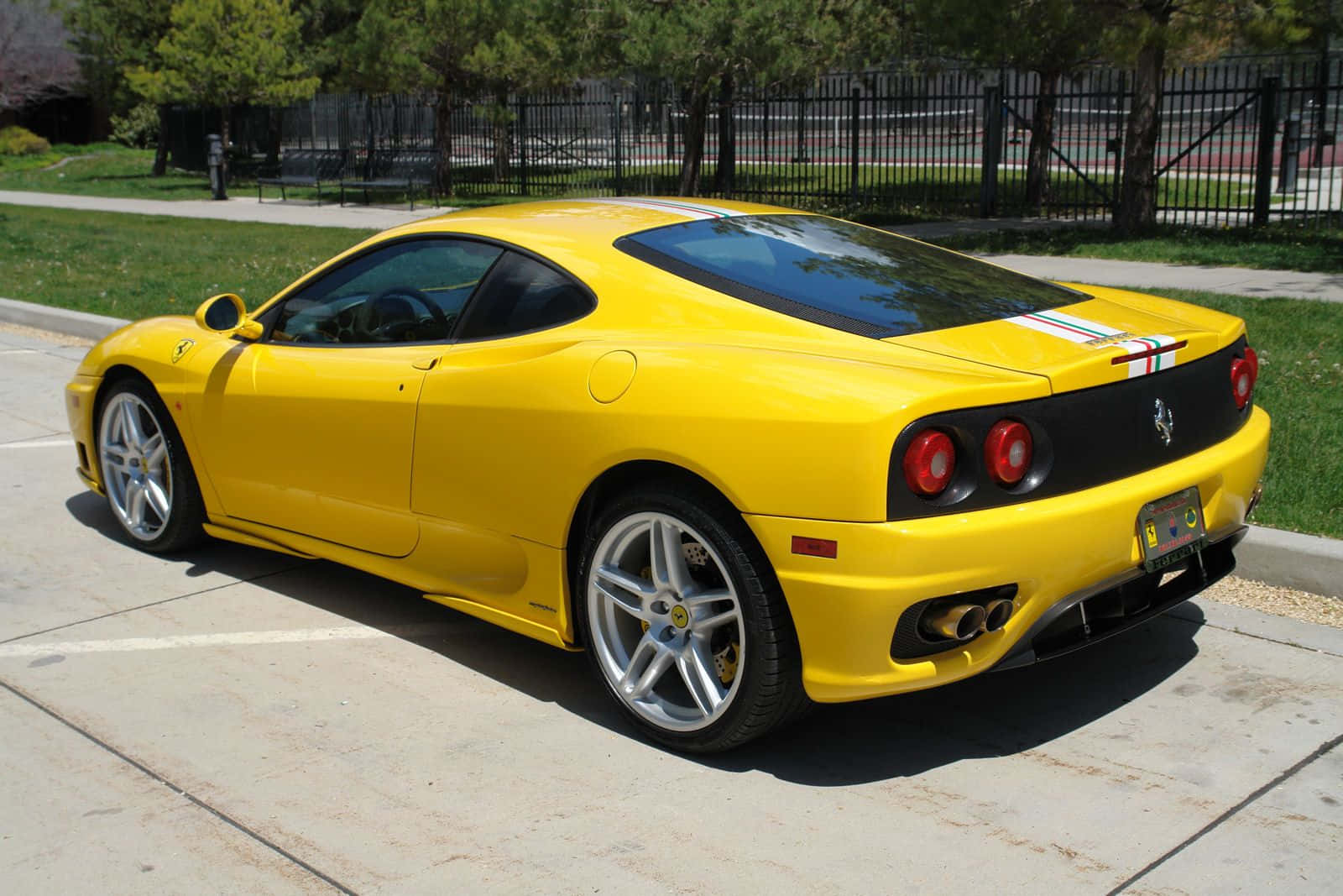 Ferrari 360 Modena 桌布 壁紙 — 一輛鮮黃色的法拉利360跑車停泊在陽光明媚的混凝土車道上，背景有綠樹和圍欄。