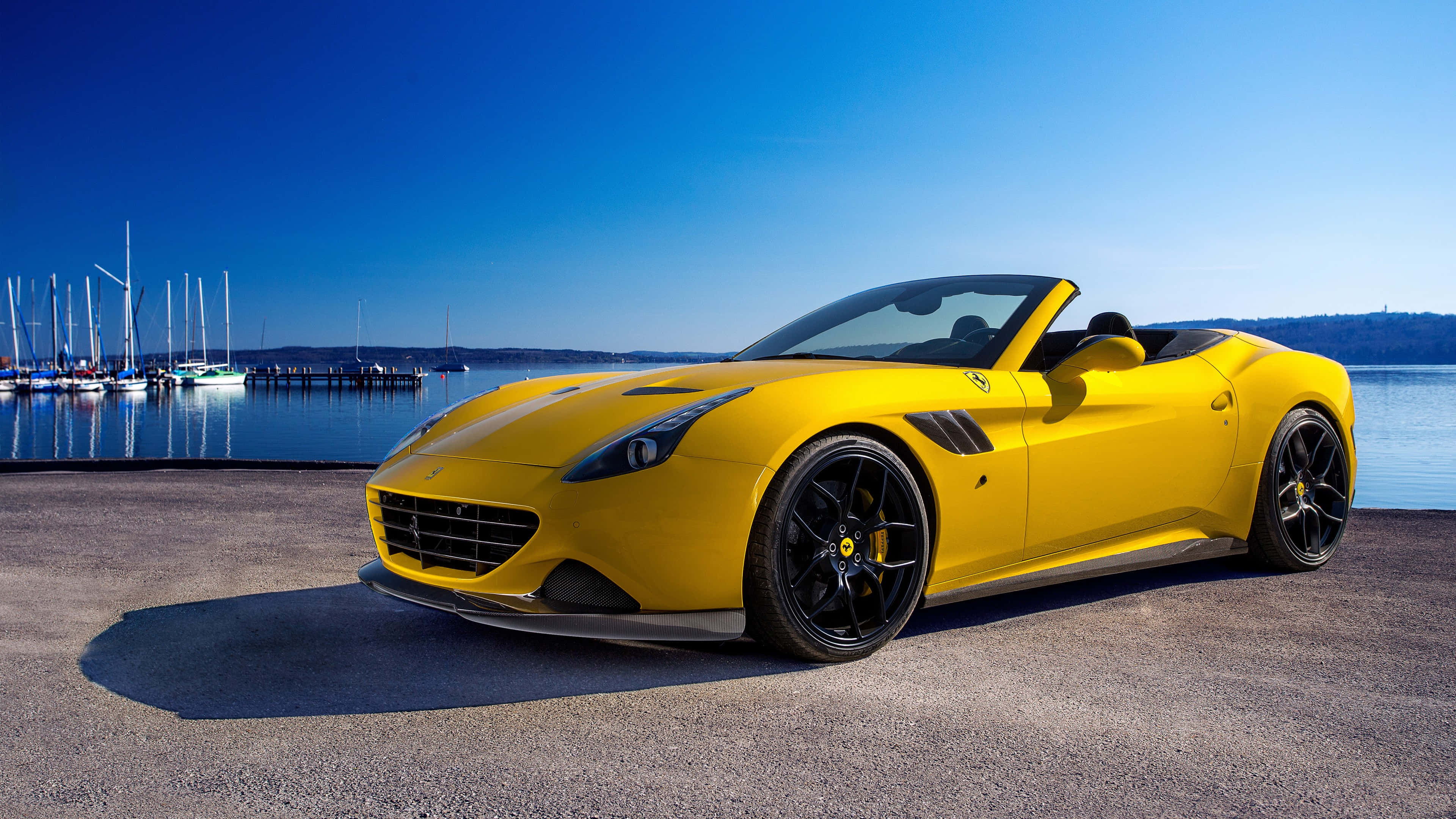 Ferrari Portofino 桌布 壁紙 — 一輛鮮黃色的法拉利開篷車停泊在碼頭上，旁邊有帆船和寧靜的水面，天空清澈湛藍。
