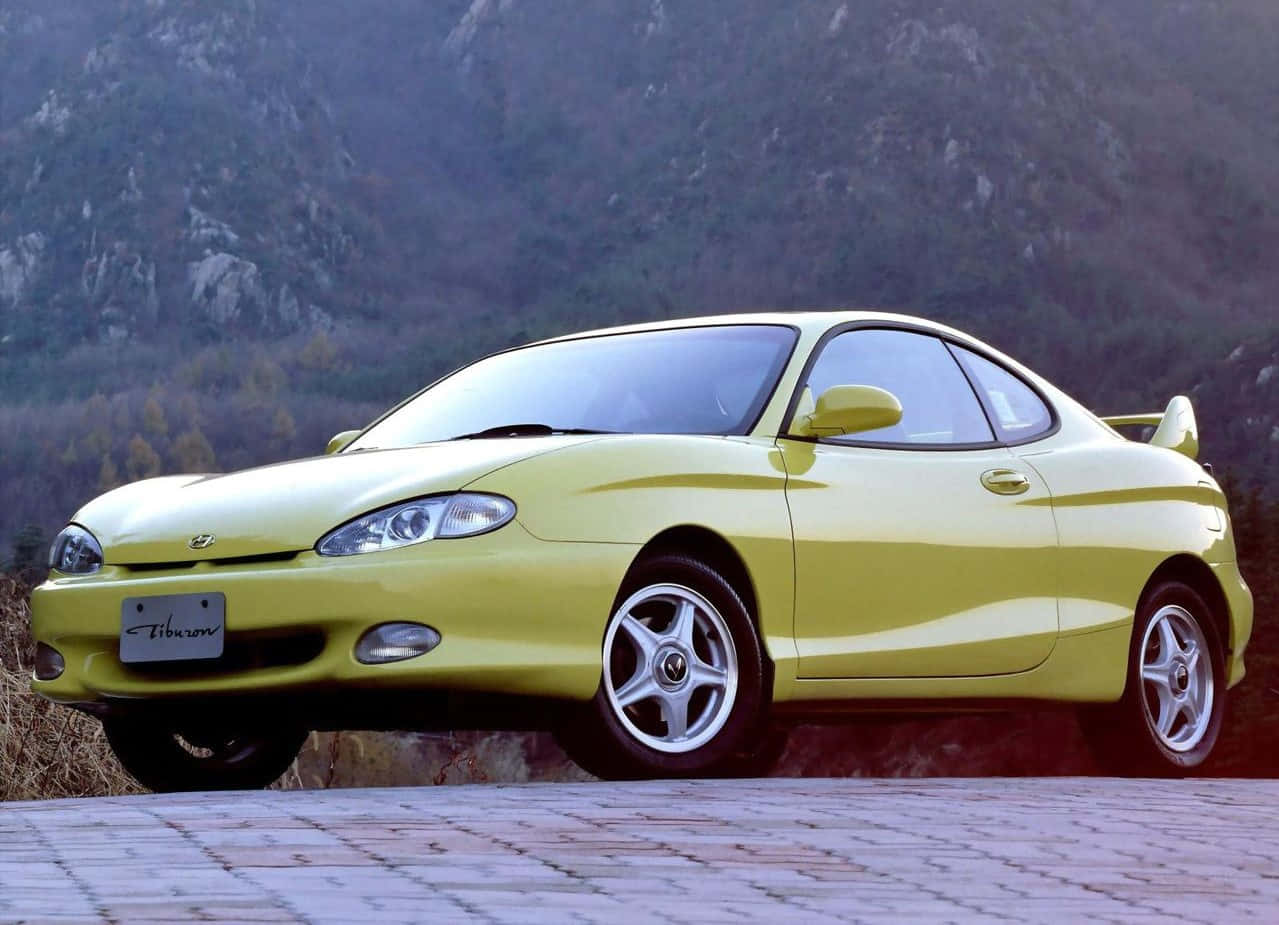 Hyundai Tiburon 桌布 壁紙 — 一輛鮮黃色的現代 Tiburon 雙門跑車停泊在石徑上，背景是岩石山脈和秋天的樹木。