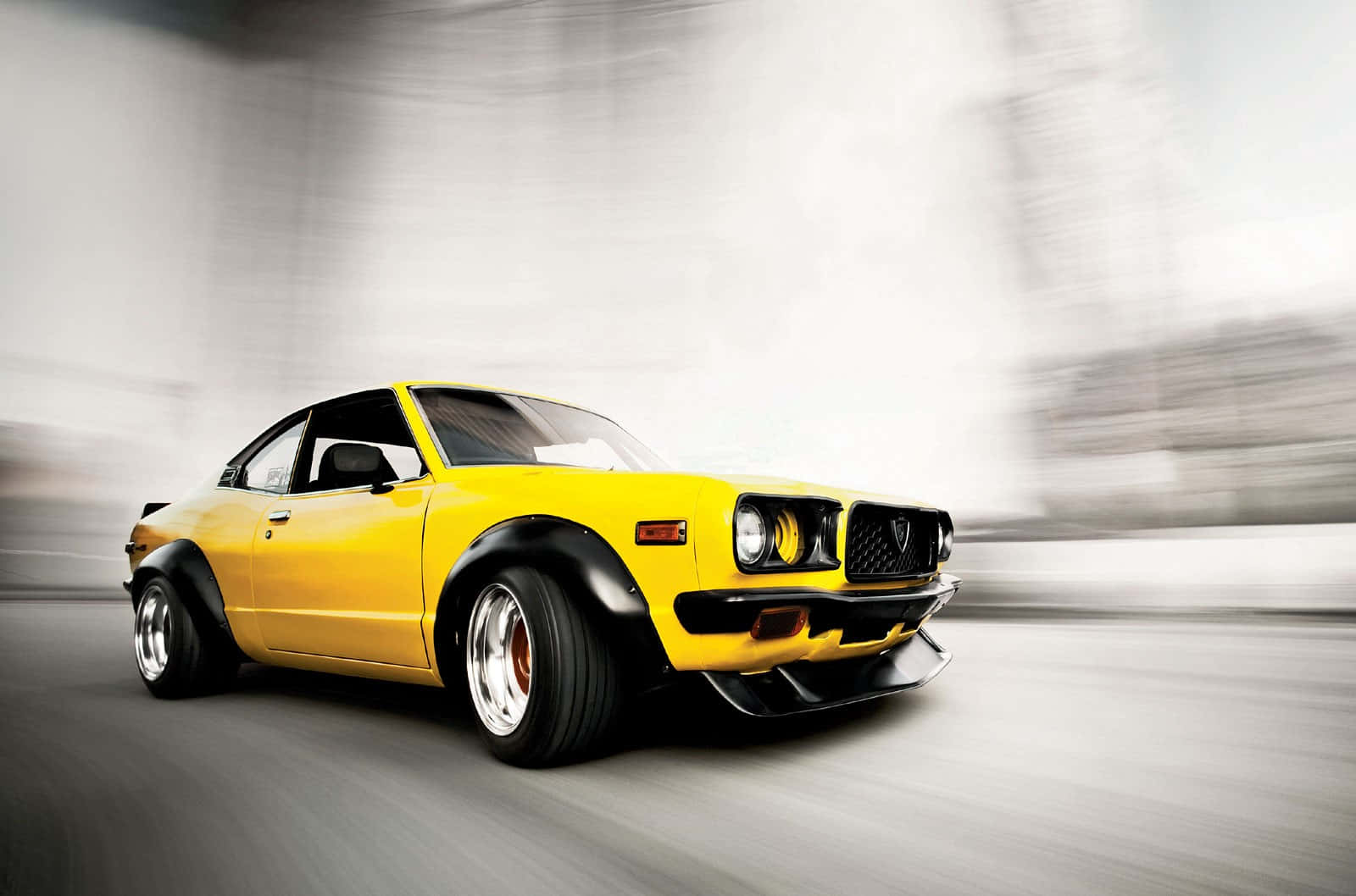 Mazda Rx-3 桌布 壁紙 — 一輛鮮黃色的經典跑車向前飛馳，動態模糊劃過灰色的城市背景。