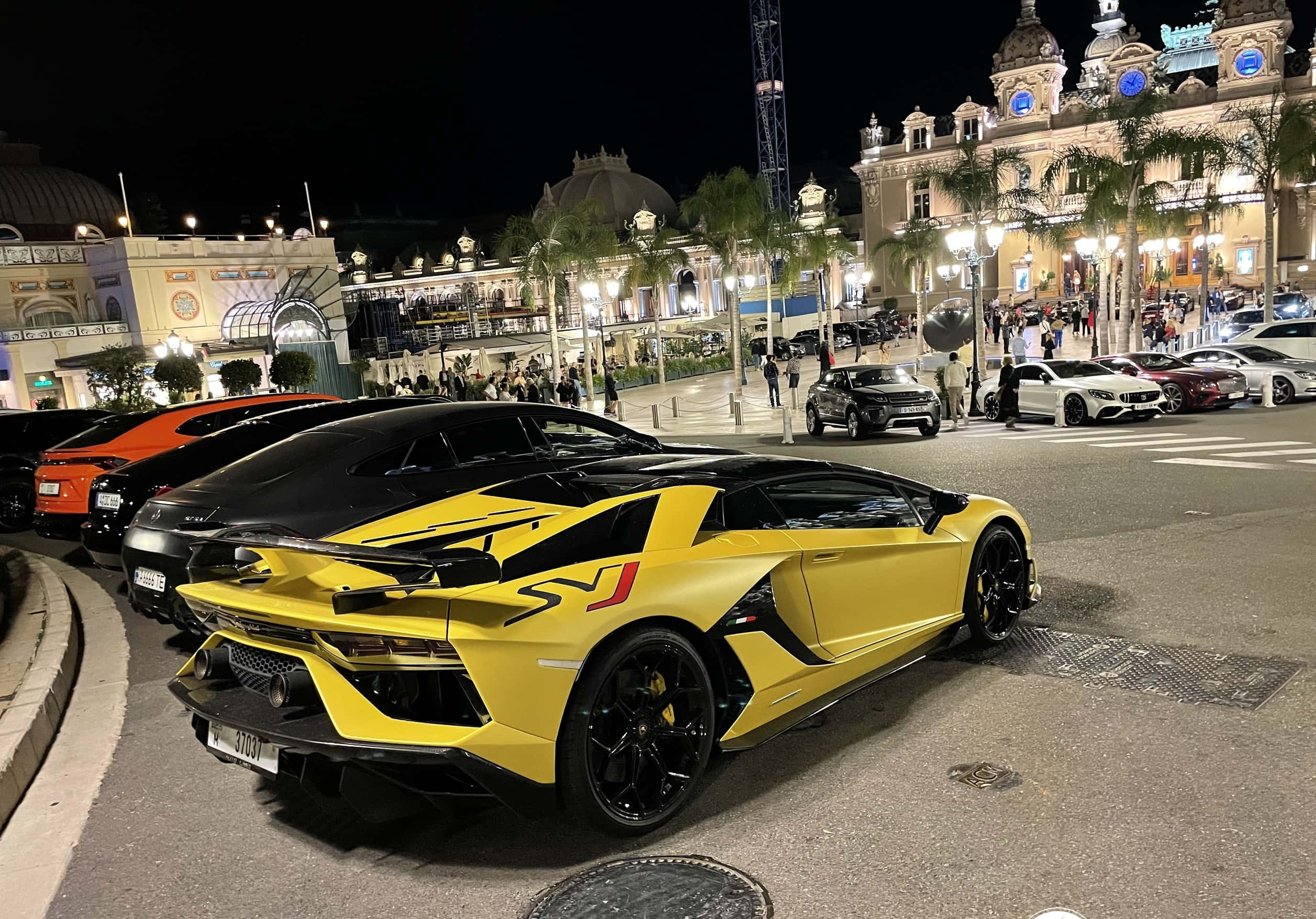 一輛黃色跑車停在建築前 壁紙 — 一輛鮮黃色的林寶堅尼 Aventador SVJ 在夜晚停泊在繁華的豪華度假村，周圍有燈光璀璨的建築物和棕櫚樹。