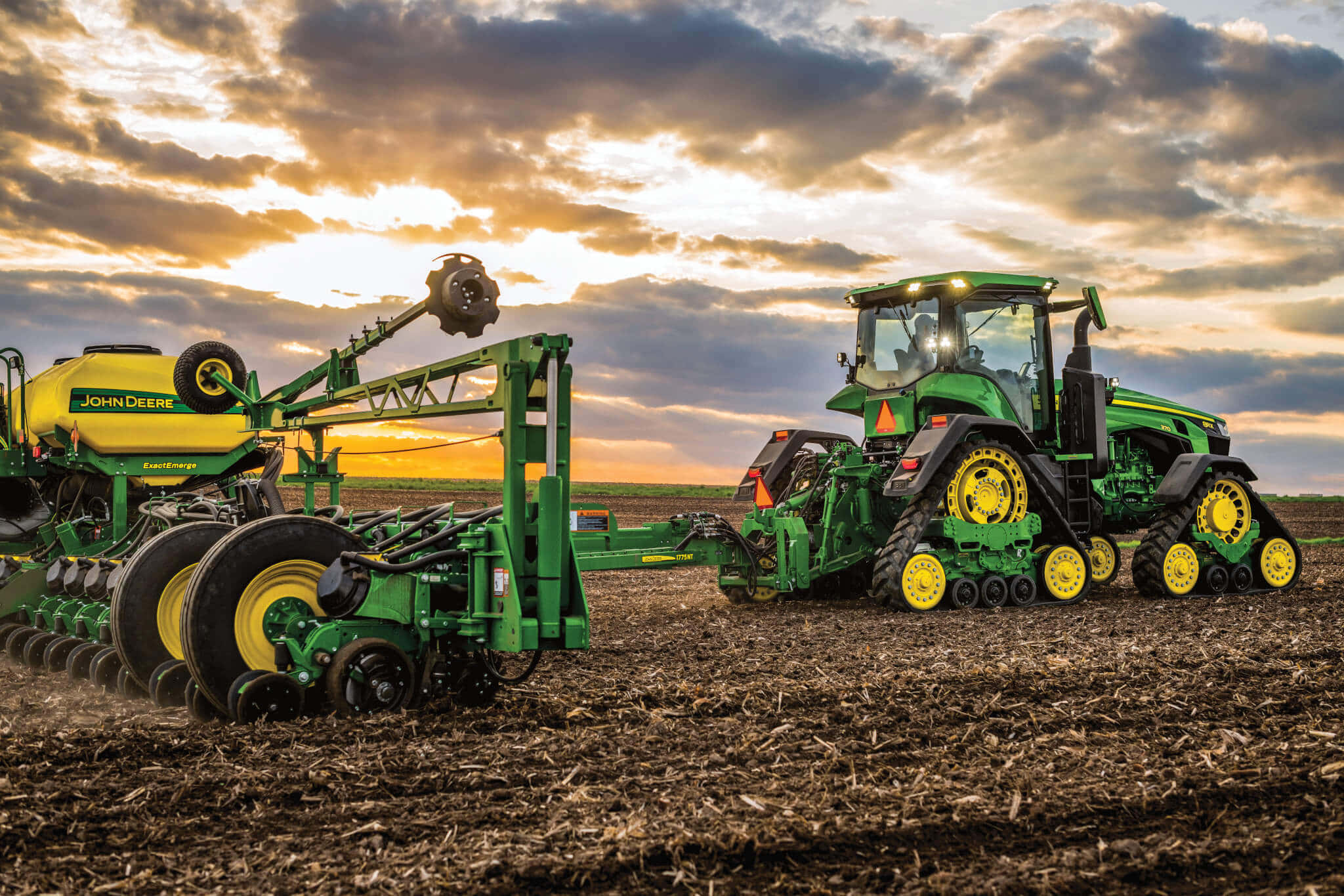 John Deere 桌布 壁紙 — 一輛黃色車輪的約翰迪爾拖拉機在日落時分，於戲劇性的雲層下拉著播種機穿過犁過的田地。