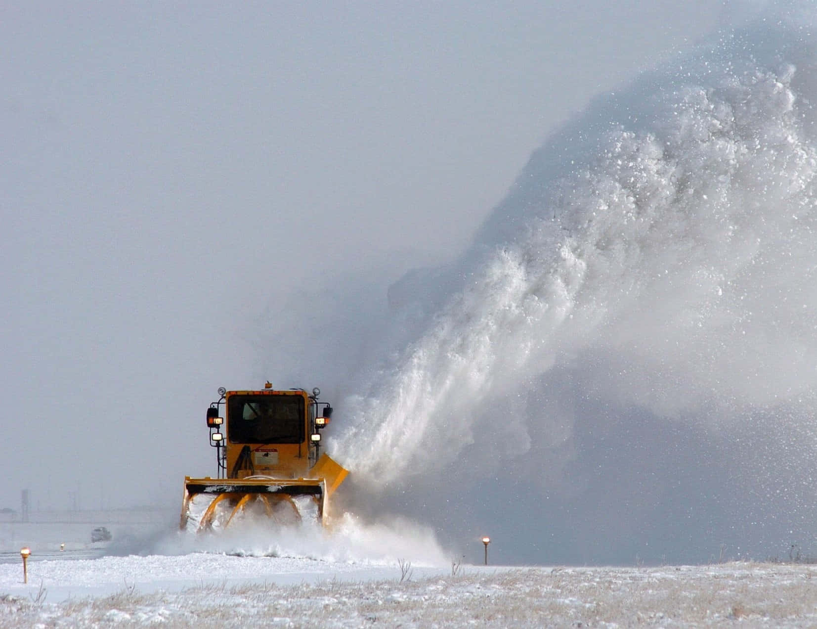 Snowplow 桌布 壁紙 — 一輛黃色除雪車在灰色冬日天空下，將巨大的雪浪吹過冰凍的田野。