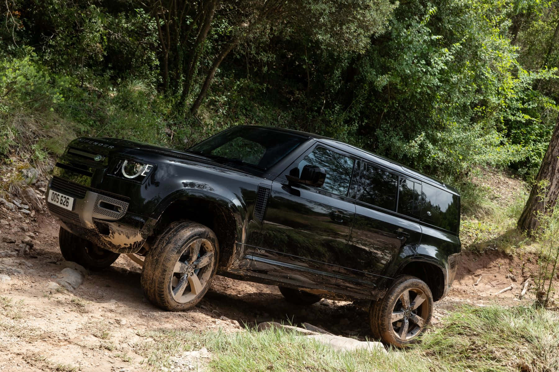 Land Rover Defender 桌布 壁紙 — 一輛黑色 Land Rover Defender 在泥濘的森林小徑上行駛，周圍是茂密的綠色樹葉。