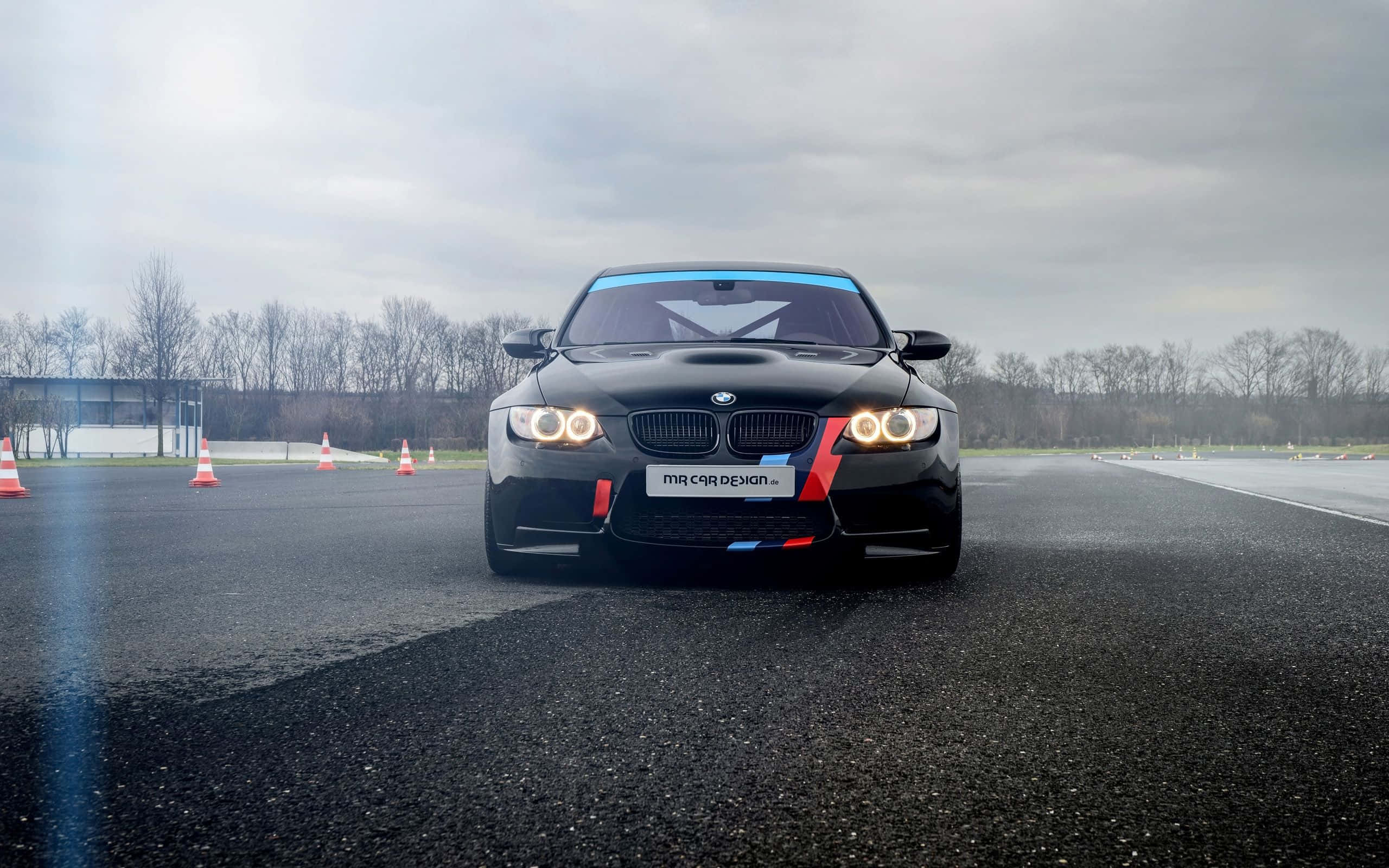 Car Design 桌布 壁紙 — 一輛黑色BMW M3賽車，車頭燈發亮，在陰天下的濕瀝青賽道上，前方擺放著橙色錐筒。