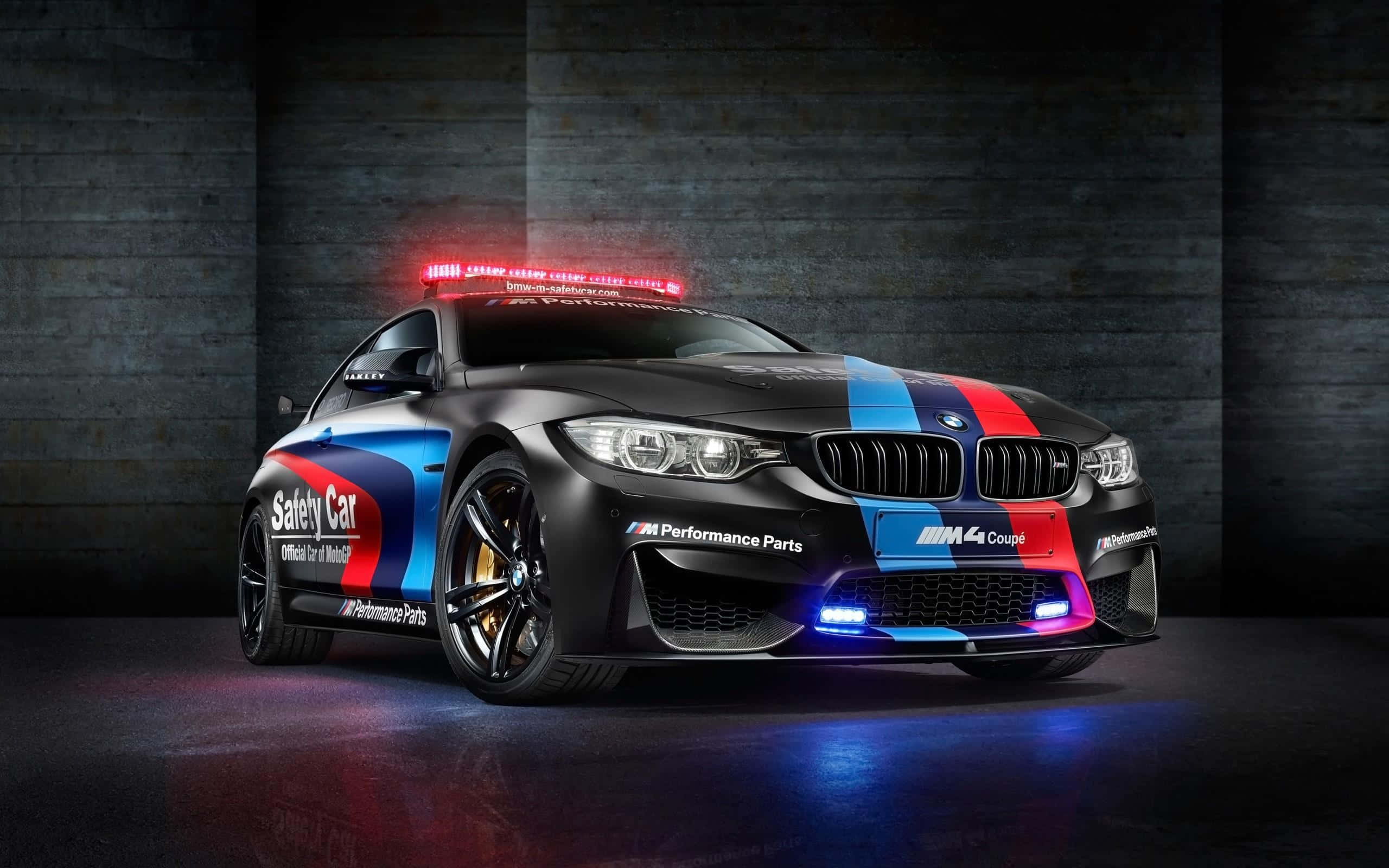 一架型格安全車在風光明媚的日子領跑賽事 壁紙 — 一輛黑色BMW M4 Coupe安全車，帶有紅、藍、白色賽車條紋，停在黑暗的混凝土車庫中，燈光閃爍。