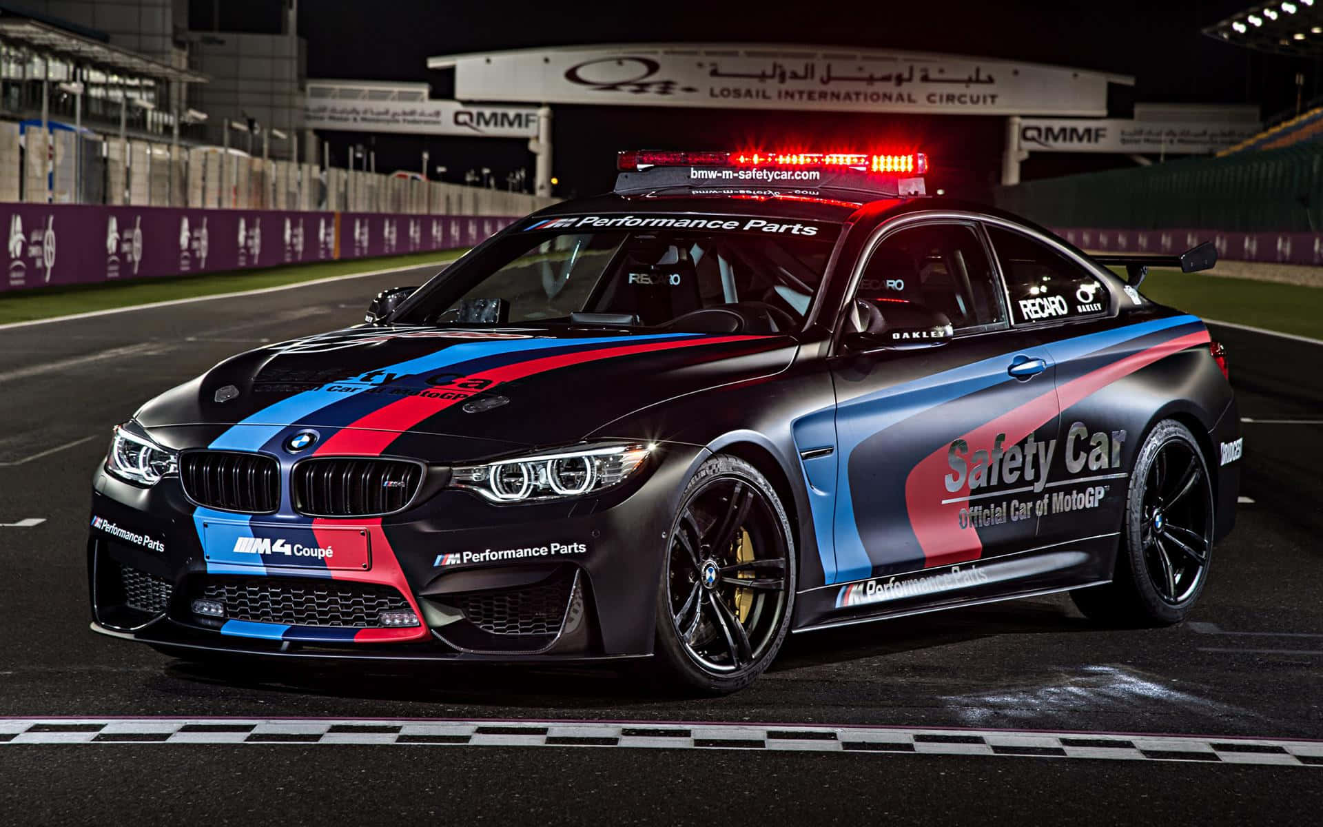 Safety Car 桌布 壁紙 — 一輛黑色BMW M4安全車，帶有紅、藍、白賽車塗裝，在夜間明亮的體育場燈光下沿著潮濕的賽道飛馳。