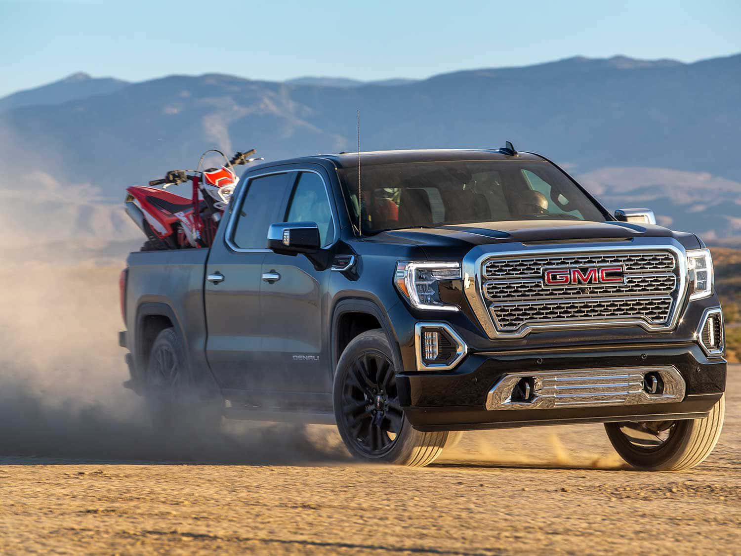 Gmc Sierra 桌布 壁紙 — 一輛黑色GMC Sierra Denali皮卡車在陽光明媚的沙漠中行駛，揚起灰塵，背景有山脈。