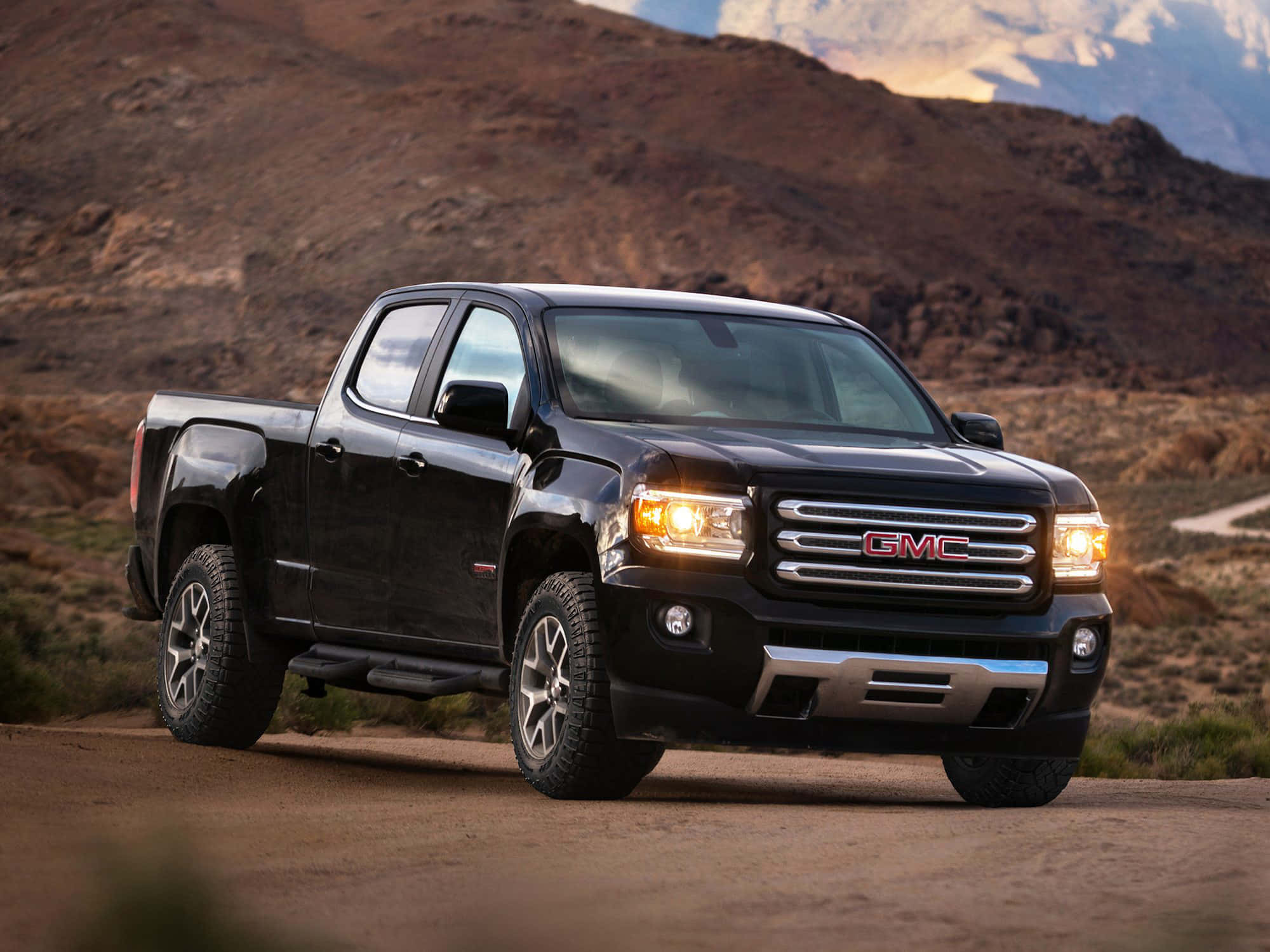 Gmc Canyon 桌布 壁紙 — 一輛黑色GMC Sierra皮卡車，車頭燈亮著，停在塵土飛揚的沙漠道路上，背景是崎嶇的山脈和日落。