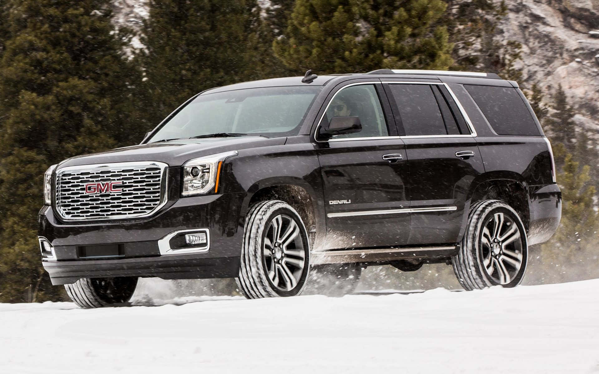 Gmc Yukon 桌布 壁紙 — 一輛黑色GMC Yukon Denali SUV在深雪中行駛，輪胎濺起雪花，背景是常綠樹和雪山。
