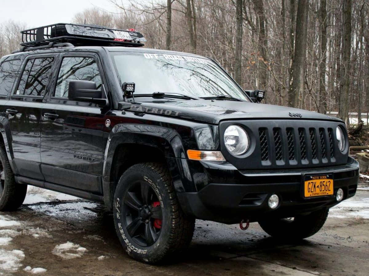 Jeep Patriot 桌布 壁紙 — 一輛黑色Jeep Patriot配備車頂行李架和越野燈，停在雪地上，附近有光禿禿的樹木。