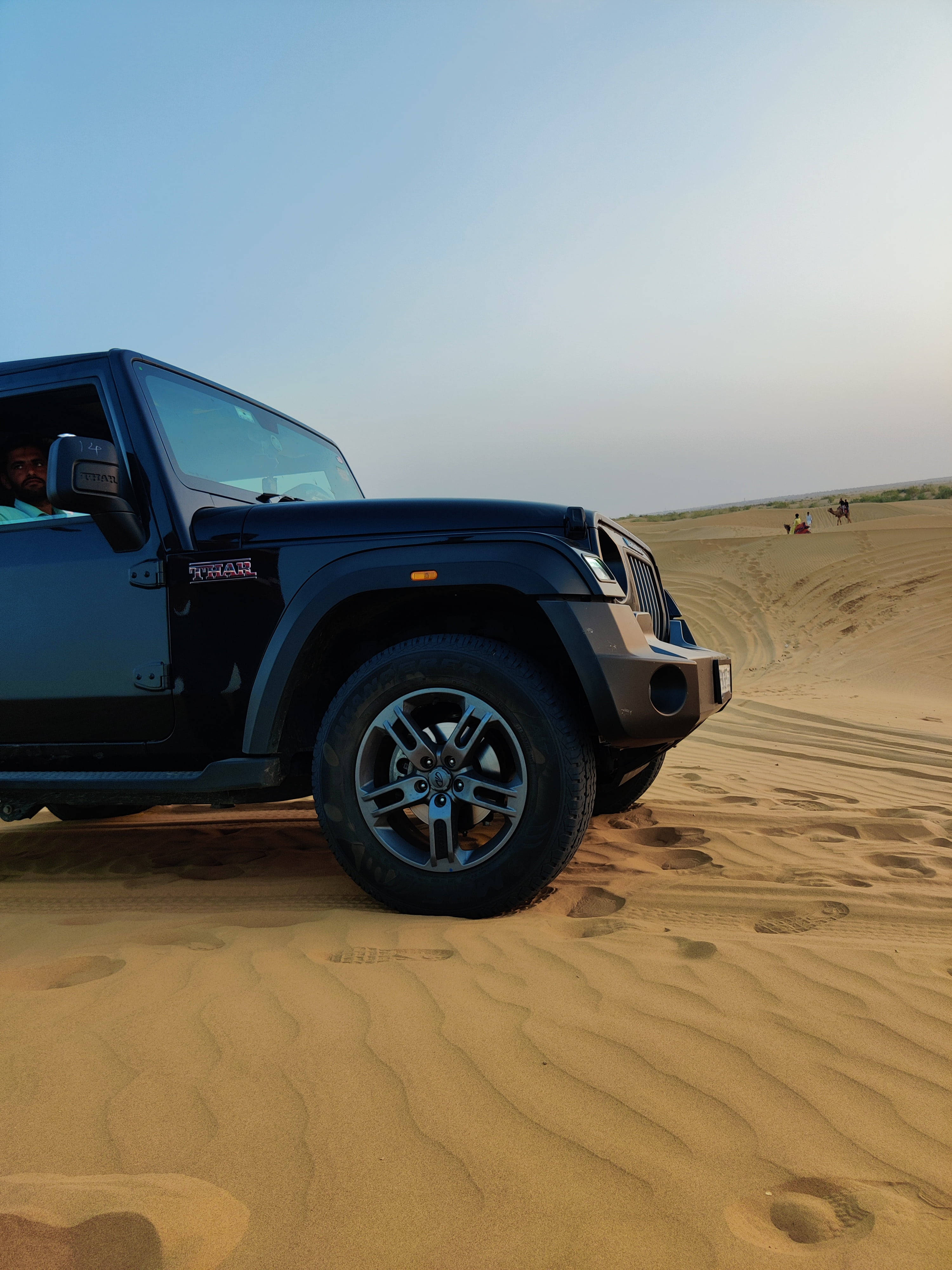 肖像 桌布 壁紙 — 一輛黑色Jeep Wrangler在晴朗藍天下駛過金色沙漠沙丘，遠處可見人影。