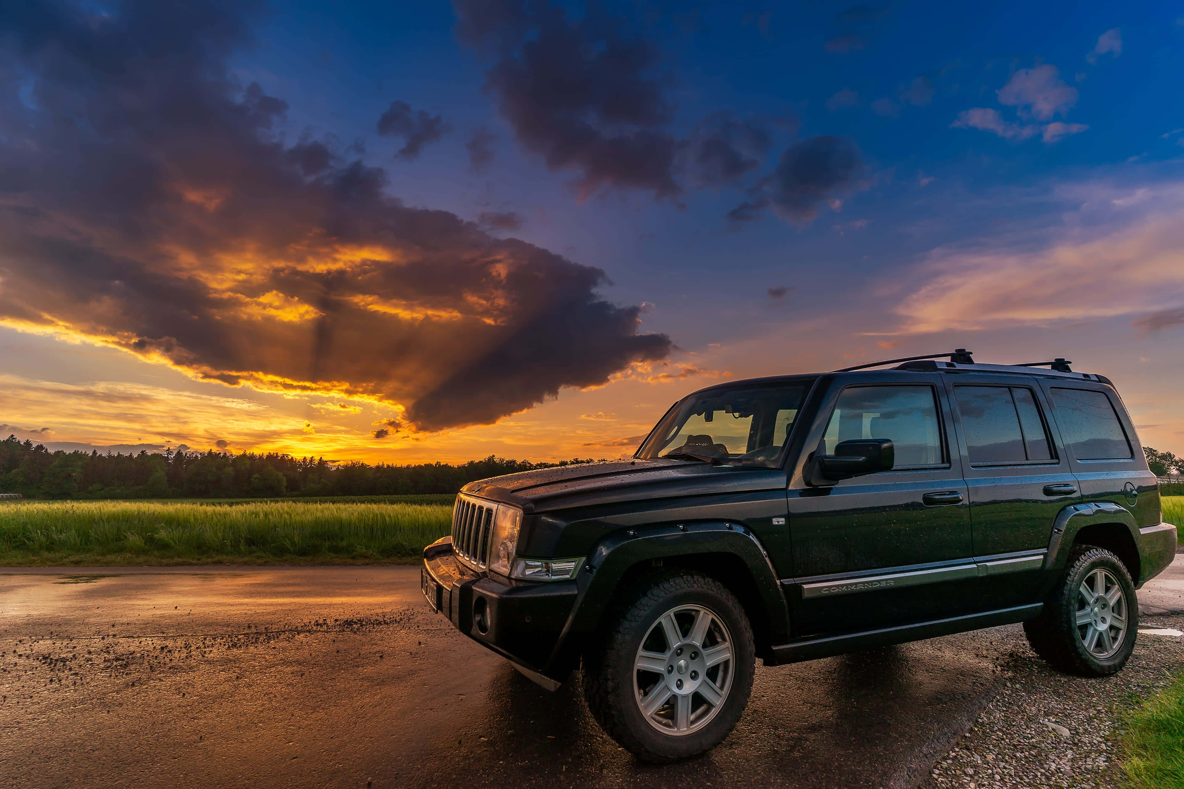Jeep Commander 桌布 壁紙 — 一輛黑色Jeep Wrangler停泊在日落時分的濕滑道路上，綠色田野上空有戲劇性的橙色和紫色雲彩。