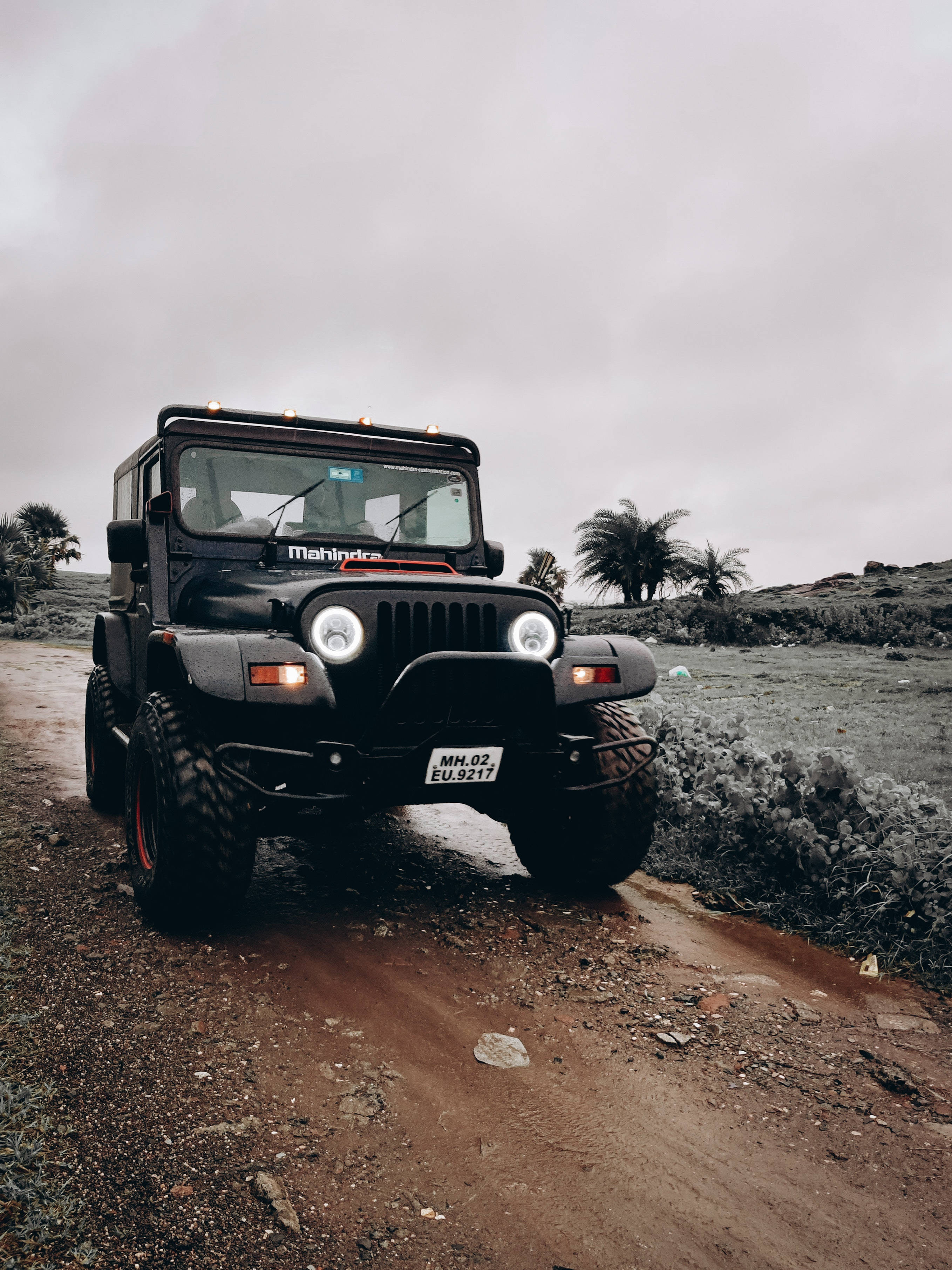 肖像 桌布 壁紙 — 一輛黑色Mahindra Thar SUV亮著車頭燈，在泥濘的越野小徑上行駛，背景是陰天和棕櫚樹。