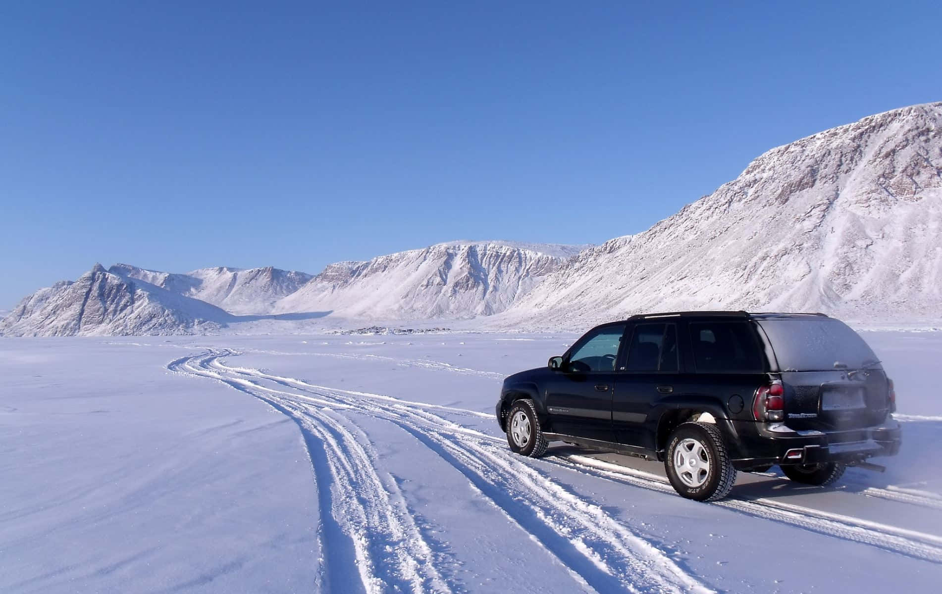 Winter Driving 桌布 壁紙 — 一輛黑色SUV在晴朗藍天下駛過廣闊的雪原，遠處是積雪覆蓋的山脈。