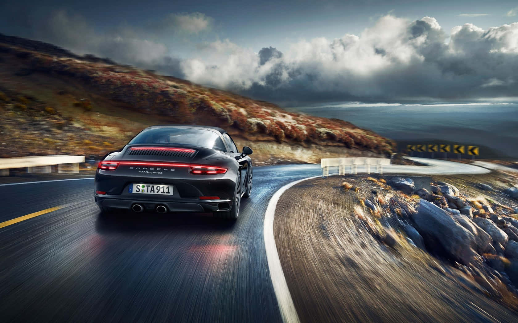 保時捷 桌布 壁紙 — 一輛黑色保時捷911 Turbo S在戲劇性的多雲天空下，沿著蜿蜒的海岸山路飛馳。