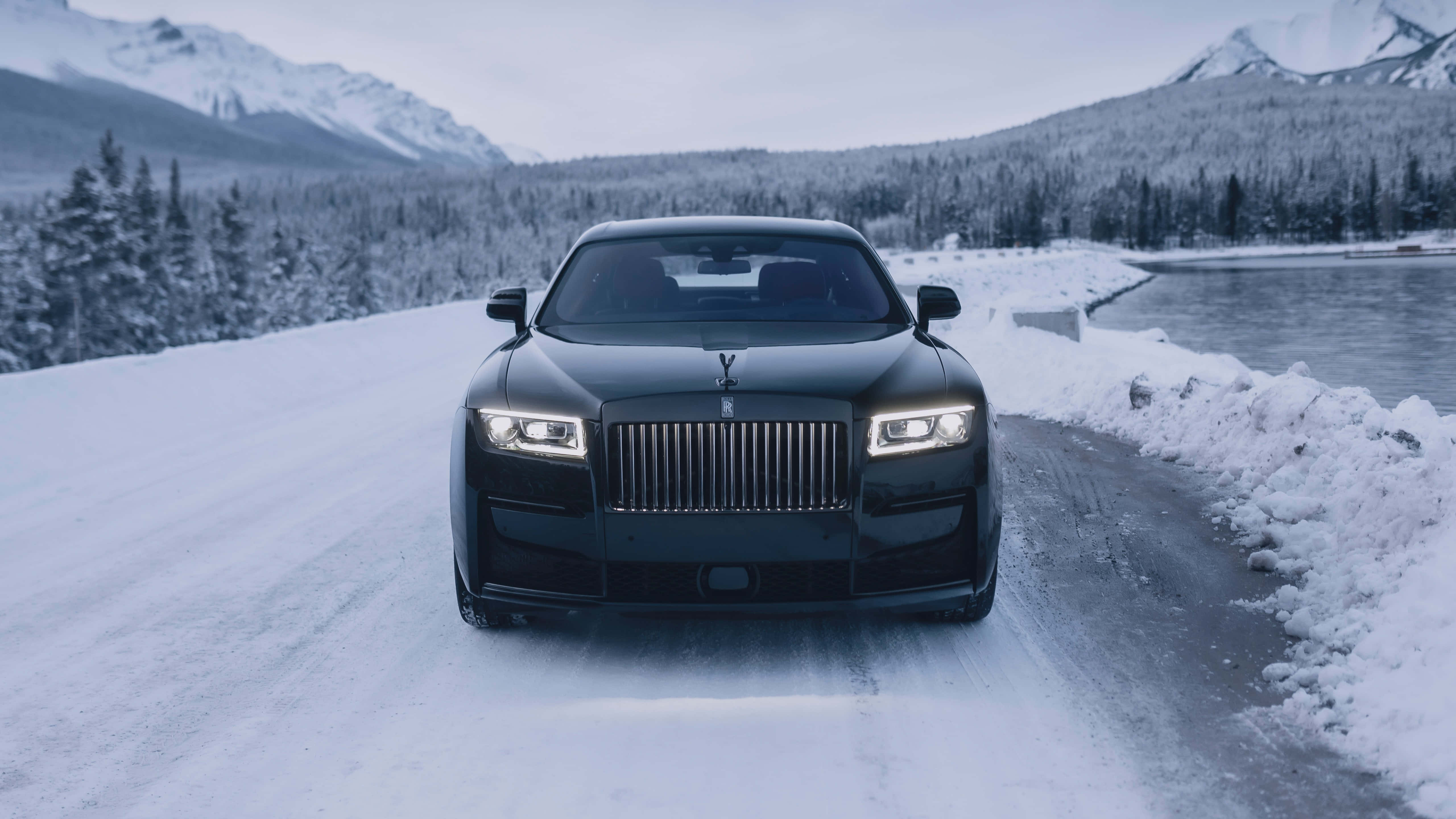 Rolls Royce 桌布 壁紙 — 一輛黑色勞斯萊斯 Ghost 在陰天行駛在結冰湖邊的積雪山路上。