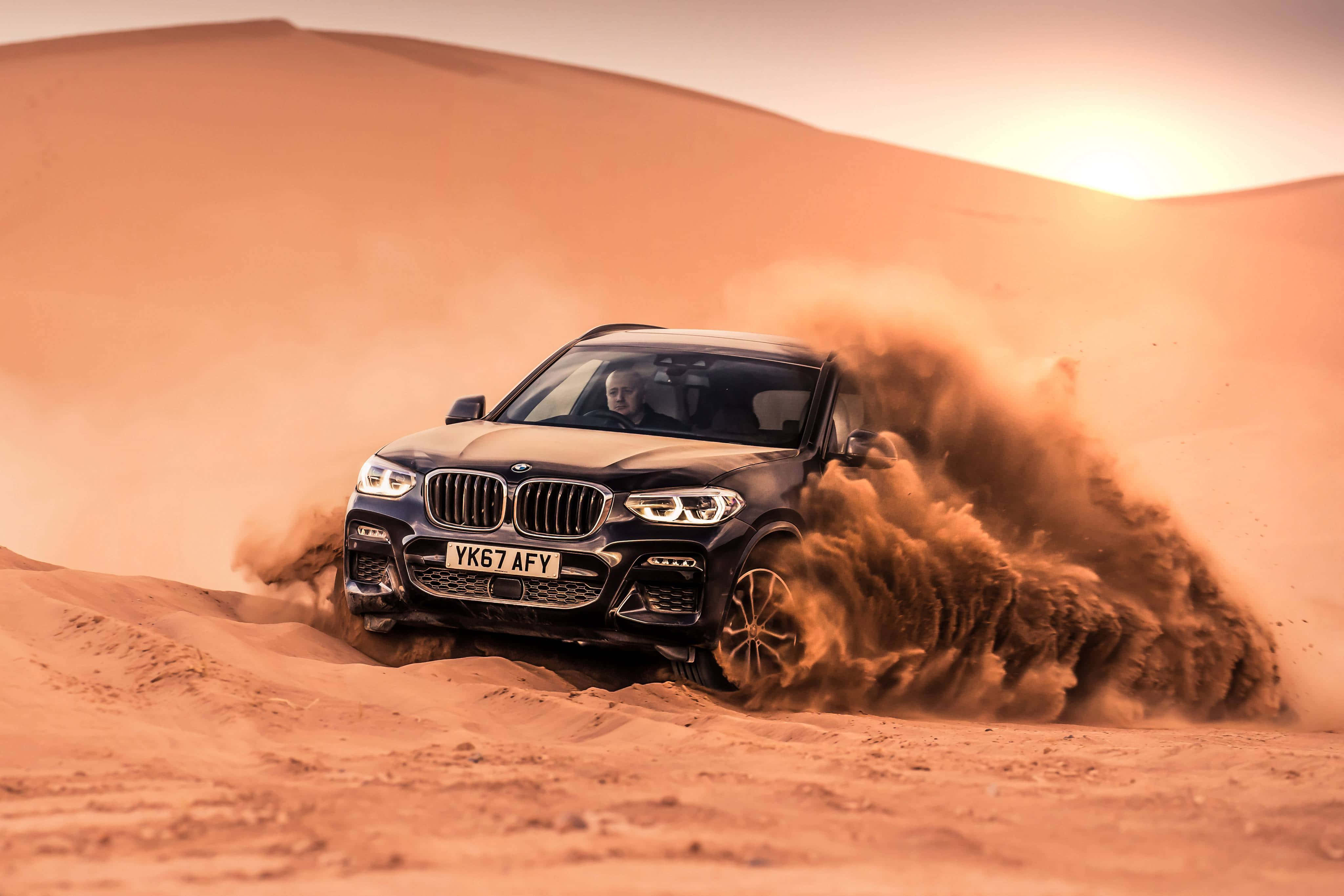 Bmw X3 桌布 壁紙 — 一輛黑色寶馬SUV在沙漠沙丘上奔馳，在溫暖的夕陽天空下揚起巨大的橙色沙塵雲。