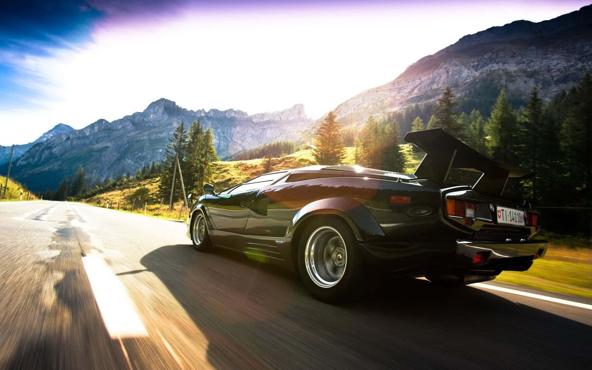 Lamborghini Countach 桌布 壁紙 — 一輛黑色林寶堅尼Countach在蜿蜒的山路上疾馳，尾翼升起，陽光照射在崎嶇的山峰和松樹林上。