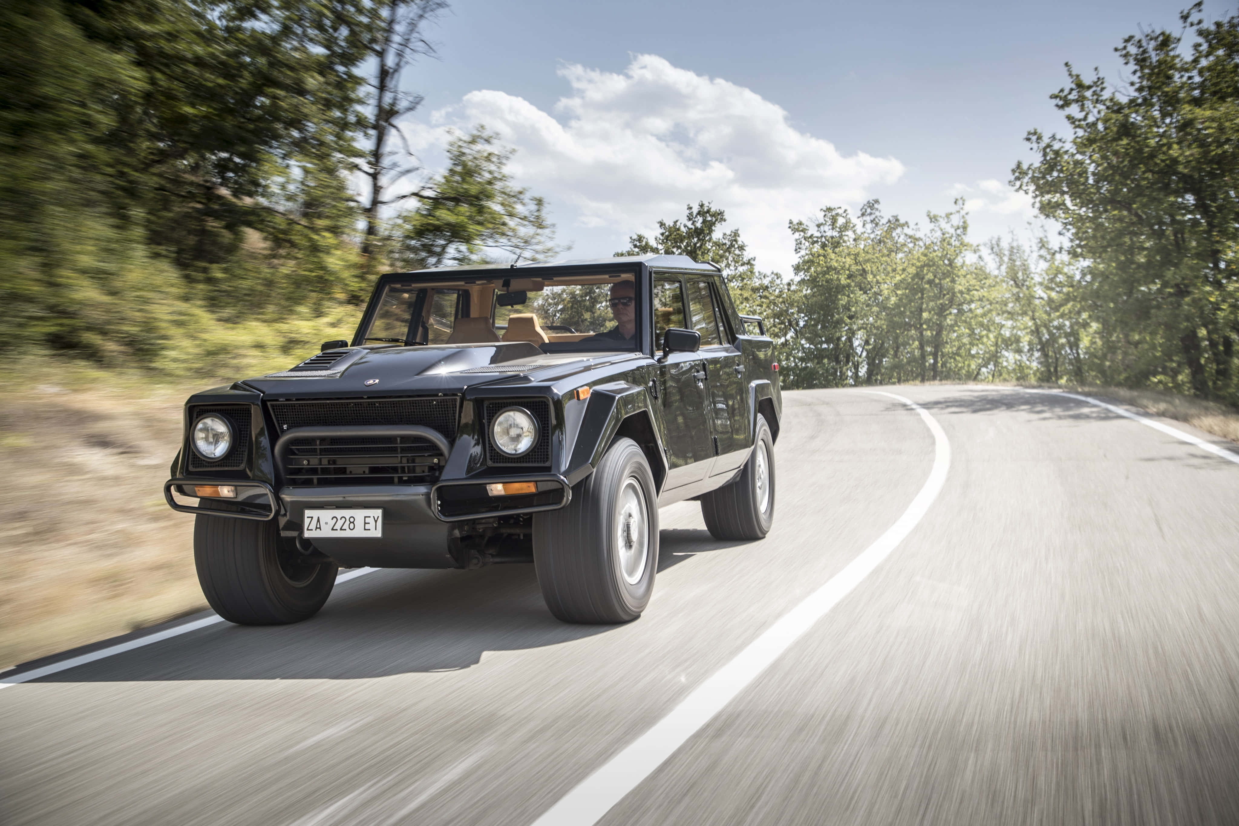 Lamborghini Lm002 桌布 壁紙 — 一輛黑色林寶堅尼LM002 SUV在晴朗藍天和蓬鬆白雲下，沿著蜿蜒的森林道路飛馳。
