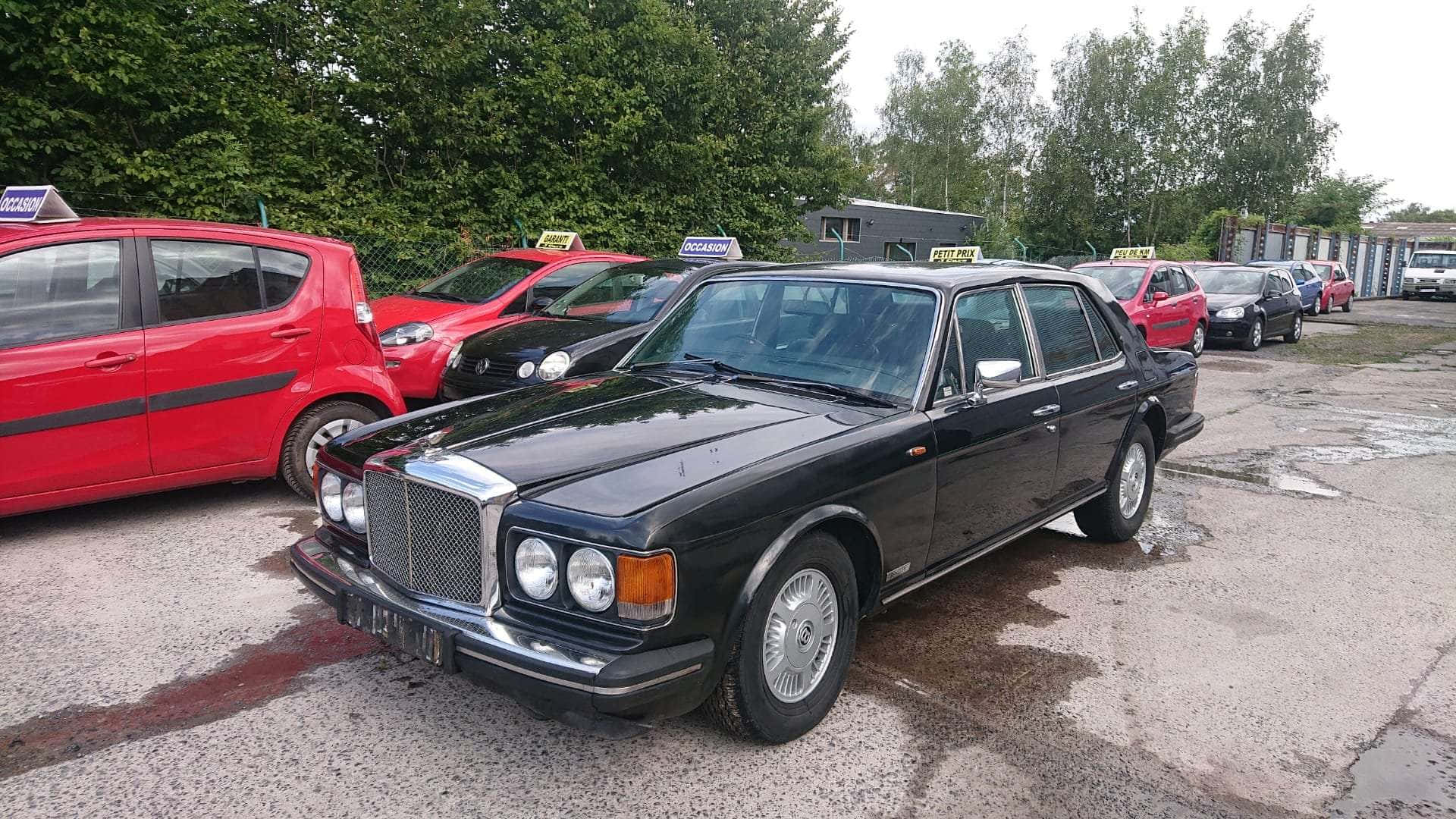 Bentley Eight 桌布 壁紙 — 一輛黑色經典賓利房車停在潮濕的停車場，周圍是色彩繽紛的的士和綠樹。