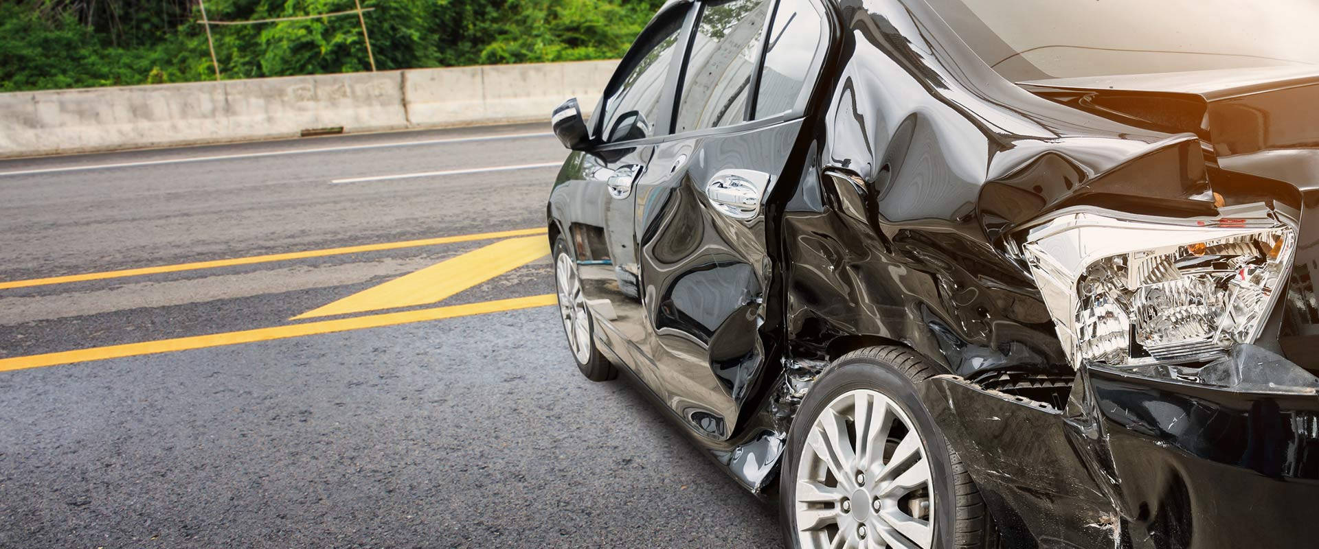 car crash 桌布 壁紙 — 一輛黑色轎車車頭嚴重受損，停在陽光明媚的道路上，旁邊是混凝土護欄和綠色植物。