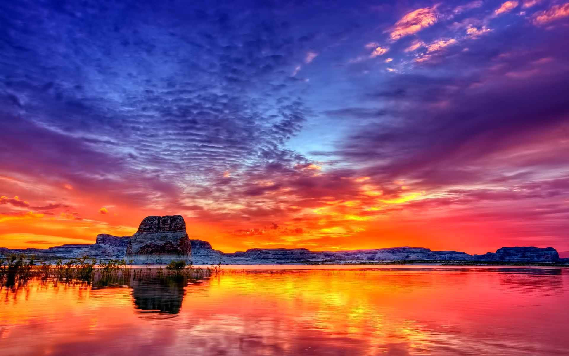 日落 桌布 壁紙 — 一輪充滿活力的夕陽將天空染成火橙色和深紫色，映照在平靜的湖面上，反射出色彩繽紛的雲彩和遠處的岩層。