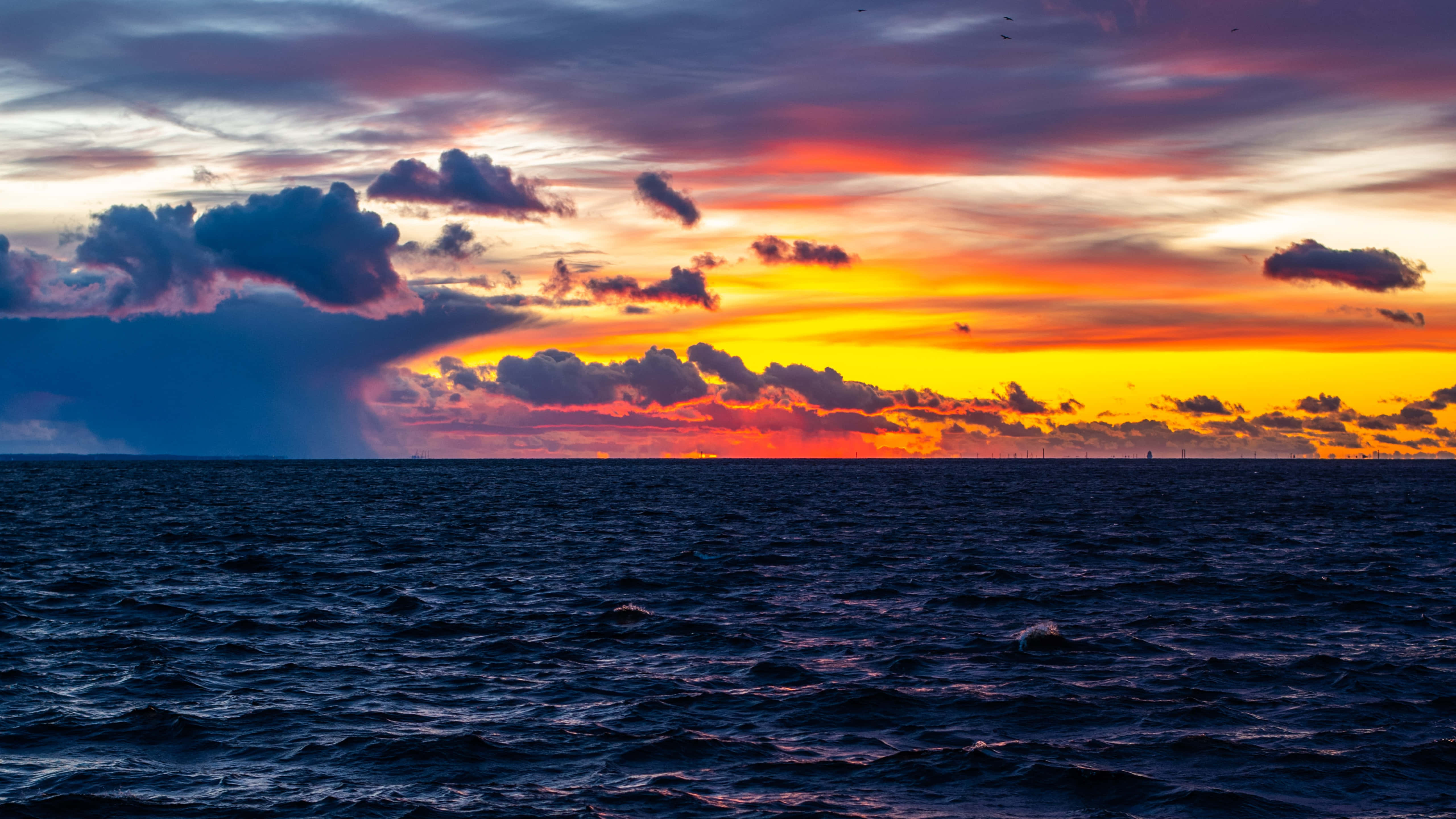 Sunset Ocean 桌布 壁紙 — 一輪戲劇性的海洋日落，火橙色和紫色的雲彩倒映在黑暗的漣漪水面上。