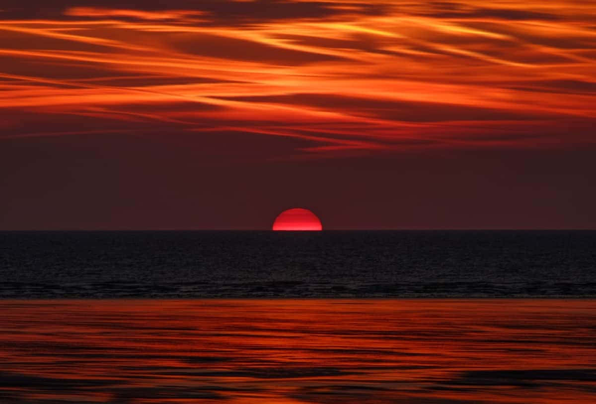 Red Sunset 桌布 壁紙 — 一輪火紅的太陽在平靜的海洋上緩緩沉入地平線，映照出鮮豔的橙色和深紅色的雲彩。