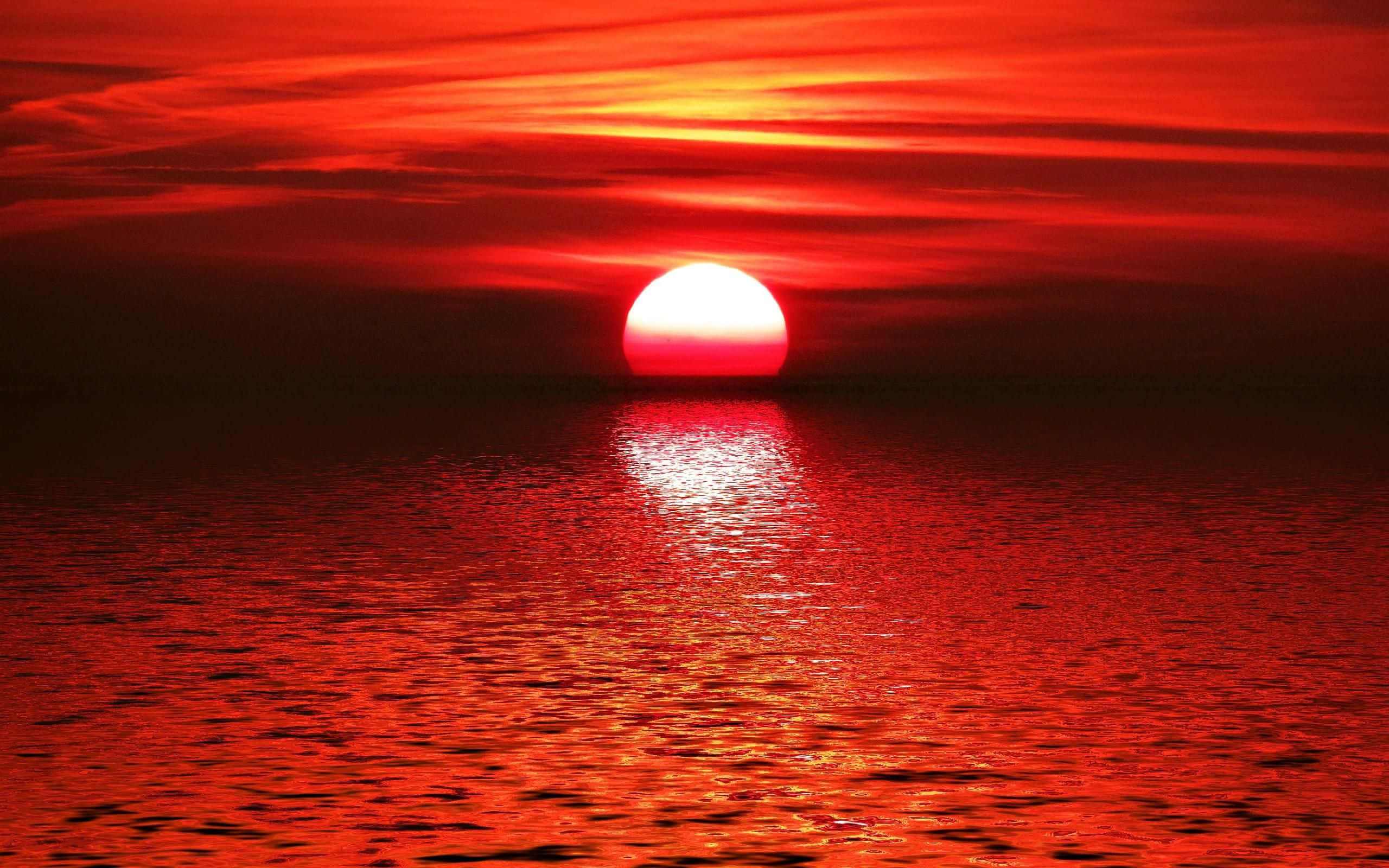 海灘日落 桌布 壁紙 — 一輪火紅的太陽在平靜的海洋上落下，在條紋雲層下於水面投射出閃爍的倒影。