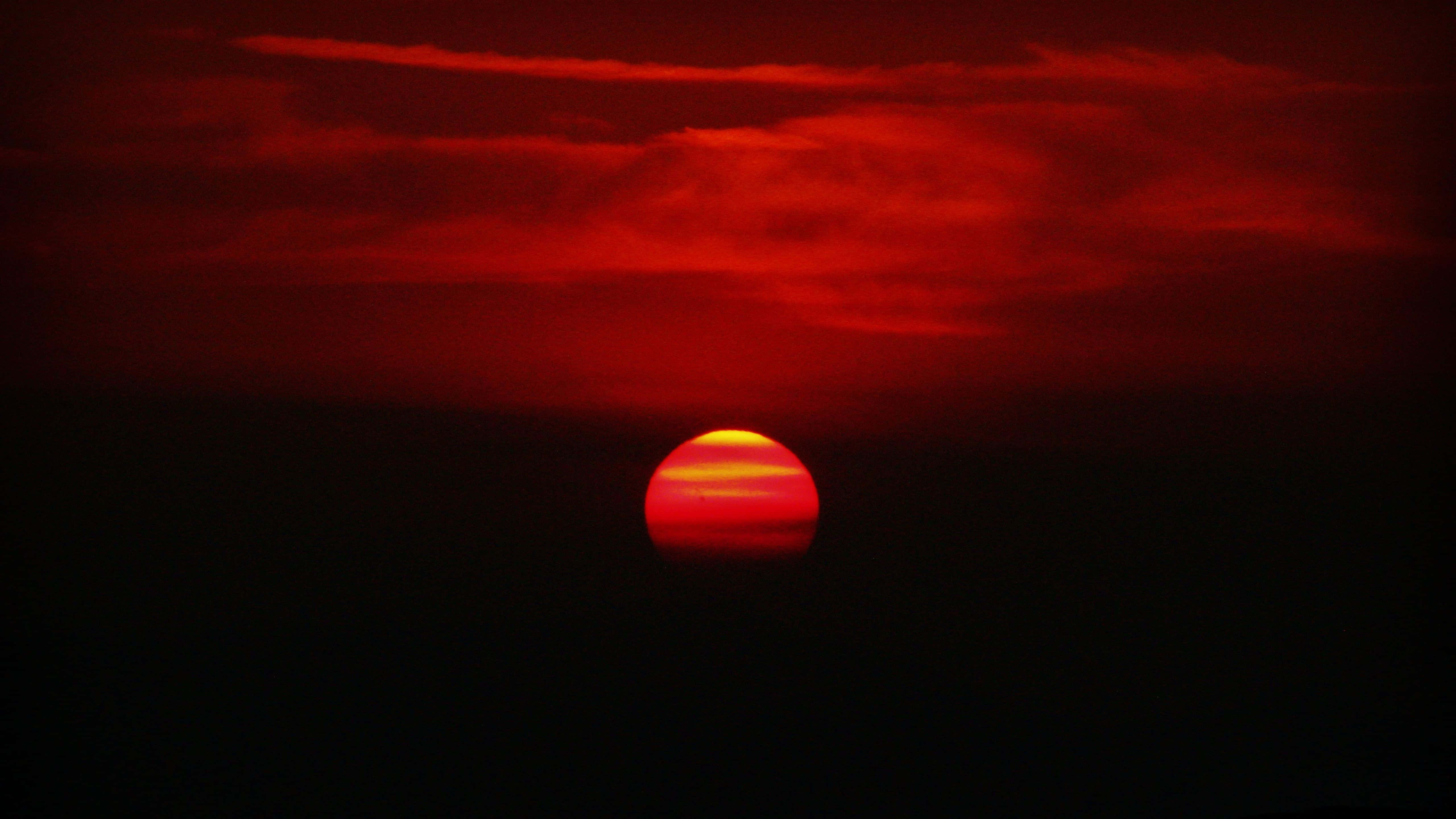 Red Sunset 桌布 壁紙 — 一輪火紅的太陽在黑暗的地平線落下，在雲層上投射出強烈的深紅色調。