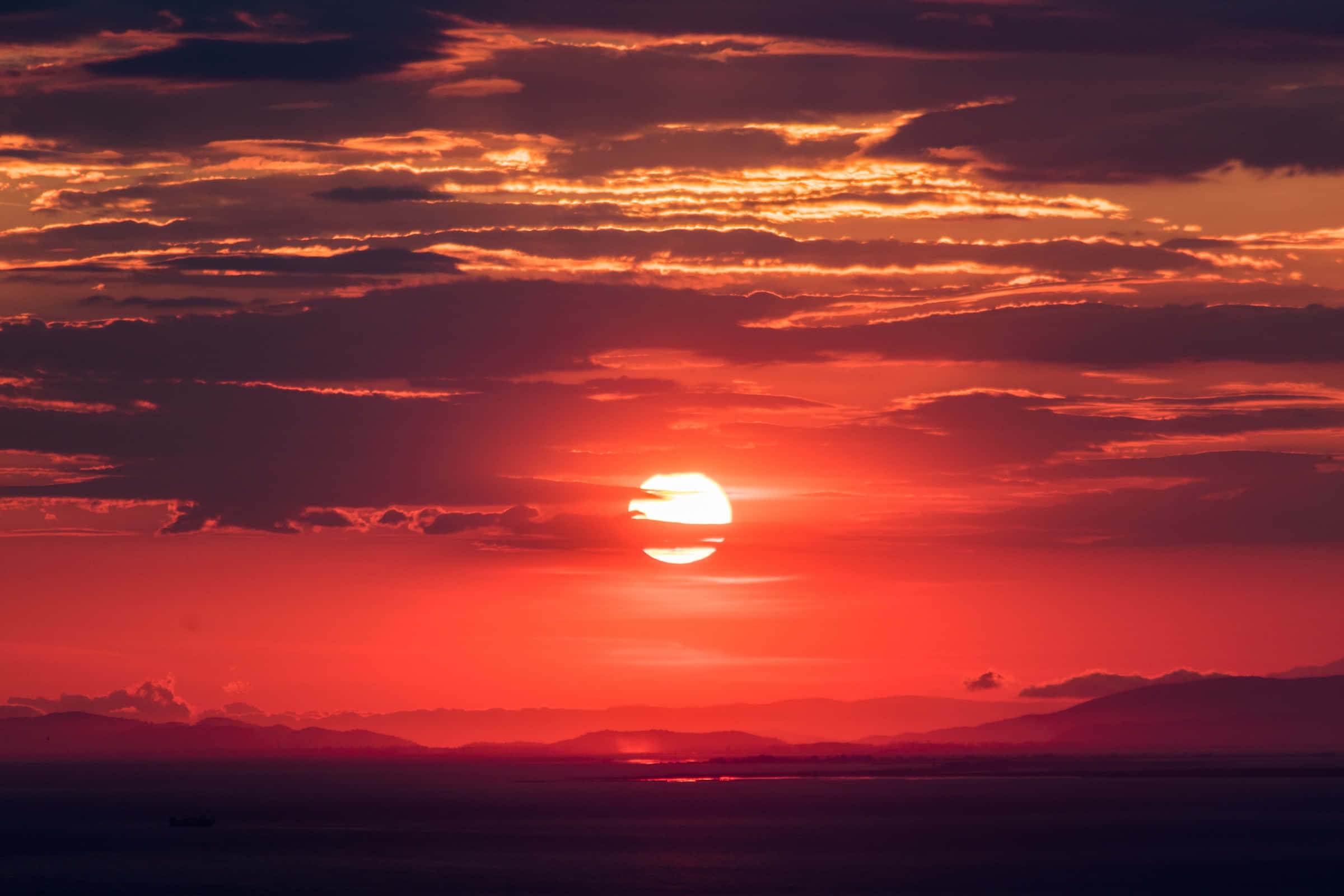 Red Sunset 桌布 壁紙 — 一輪火紅的太陽落在遠山之後，在天空和海洋上投射出鮮豔的橙色和深紅色調。