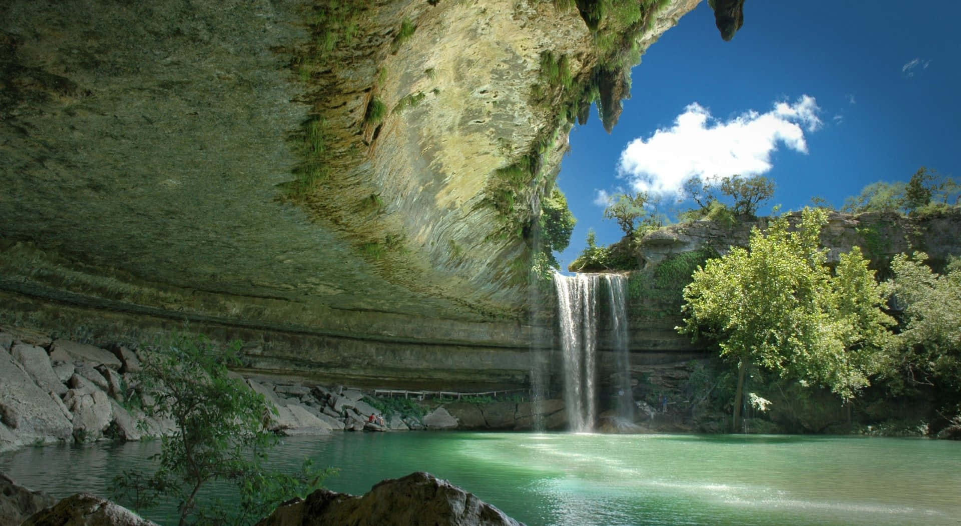 Waterfalls 桌布 壁紙 — 一道寧靜的瀑布流入一個碧綠色的水池，下方是巨大的懸崖，周圍有茂密的綠色植物，上方是明亮的藍天。